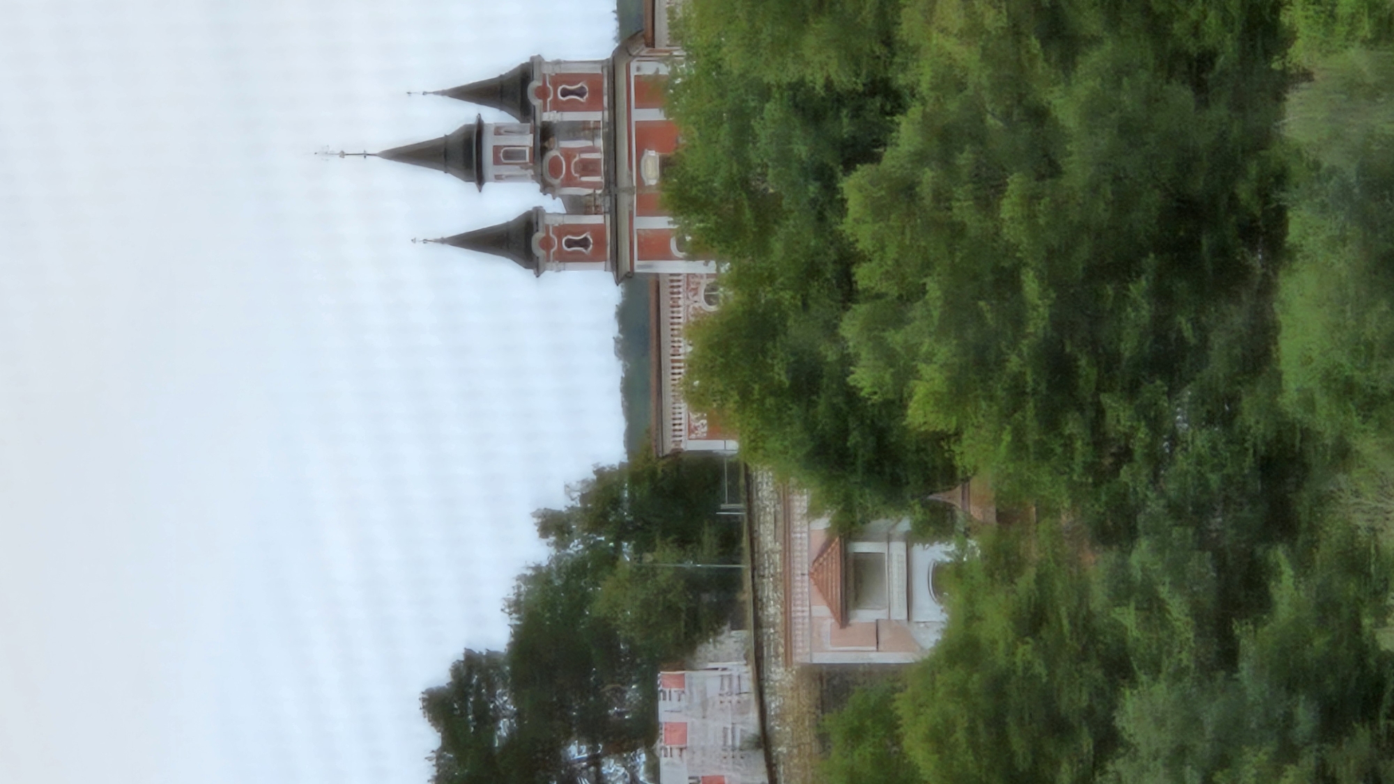 The image shows a view of the Orthodox Church of the Holy Spirit in Prešov, Slovakia. The church, with its distinct red and white facade and multiple spires, is situated on a hill and partially obscured by lush green trees in the foreground. The sky is overcast and a pale blue. To the left of the church, there appears to be construction or scaffolding on a building. The overall scene suggests a slightly overcast day with ample greenery surrounding the historical structure.