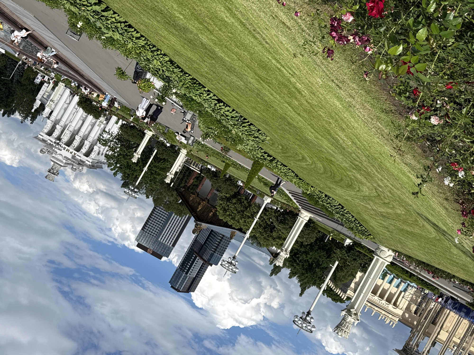 This media file depicts a sunny daytime scene in a park-like setting, likely in Moscow, Russia, given the context. The image is tilted, but shows a large expanse of manicured green grass. Along the edge of the grass is a neatly trimmed hedge. Beyond the hedge, there are several architectural structures, including a large white building with classical columns and ornate details, possibly a pavilion or exhibition hall. There are also modern high-rise buildings visible in the background, suggesting an urban park.
In the foreground and midground, there are various people milling about. Some are walking along pathways, while others appear to be sitting or standing near the buildings. The people are dressed in casual, summer clothing, indicating warm weather. There are also decorative elements such as lampposts with multiple lights and rose bushes with red and pink flowers blooming along the edge of the grassy area. The sky is blue with scattered white clouds.
Visible text includes "ОЗБЕКИСТОН" (Uzbekistan) on one of the buildings, identifying it as the Uzbekistan pavilion. The overall impression is a pleasant, leisurely day spent outdoors in a well-maintained public space.