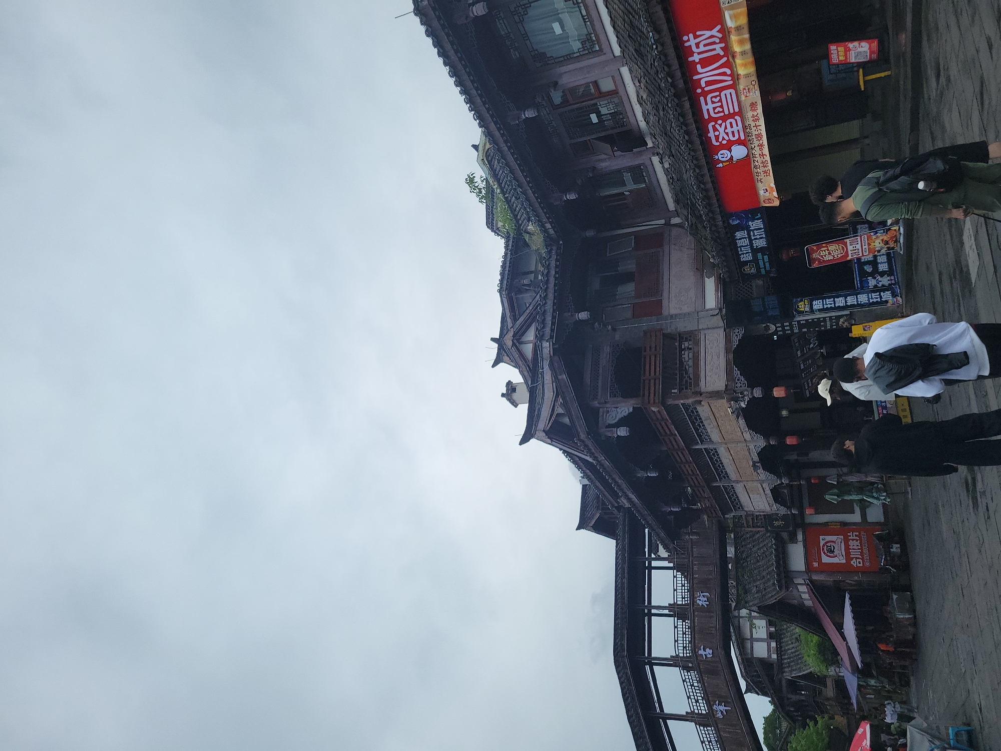 The image shows a street scene in Hechuan, China, with traditional-style architecture. The buildings are multi-storied with dark wood detailing and tiled roofs. People are walking along the street, some wearing casual clothing. There are signs in Chinese characters on the buildings, including advertisements for "蜜雪冰城" (Mixue Ice Cream & Tea) and "合川桃片" (Hechuan Peach Slice). The sky is overcast and grey. A wooden bridge structure is visible, and various shops and businesses line the street.