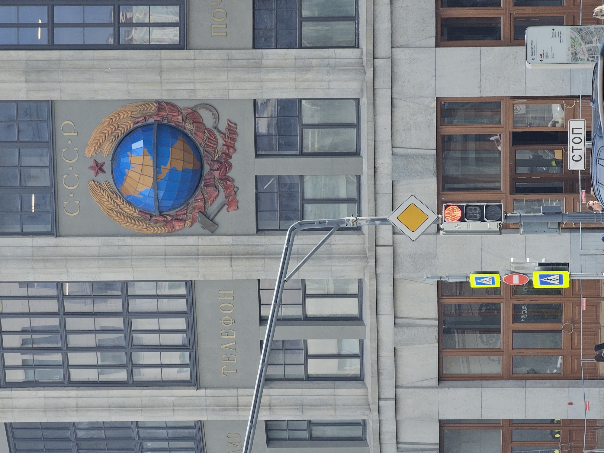 Here is a description of the image:
The image shows a section of a building facade in Moscow, Russia. Prominently displayed is the Soviet coat of arms, featuring a globe with surrounding wheat and a hammer and sickle. The Cyrillic letters "C-C-C-P" (USSR) are also visible. Gold lettering below the coat of arms spells out "ТЕЛЕФОН" (Telephone). The building features a series of windows. Street signs and traffic lights are also visible on the right. The image includes street signs written in Cyrillic, identifying the area as Tverskaya Street.