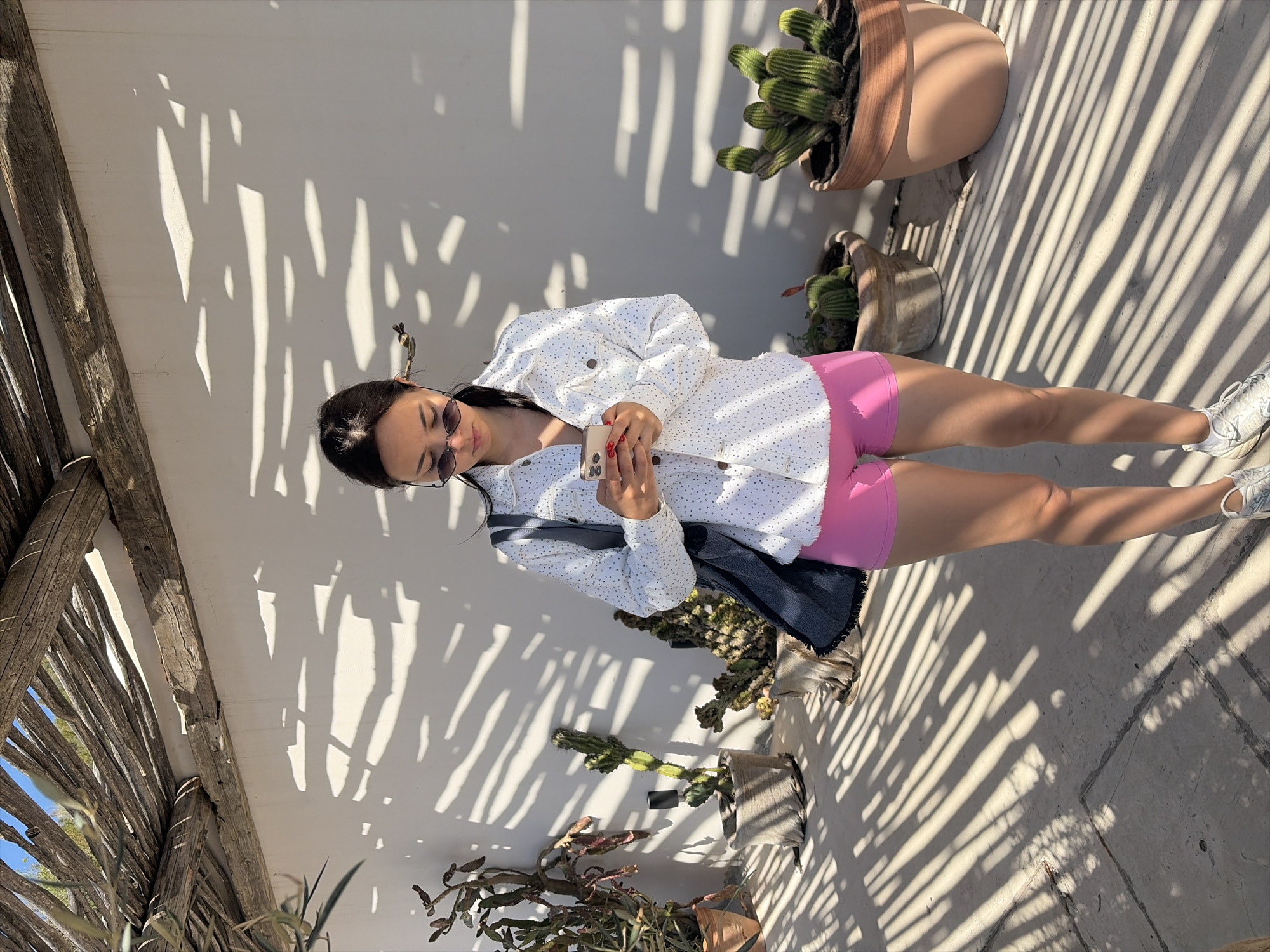 A woman with dark hair and sunglasses is standing outside in an area with dappled sunlight. She is wearing a white jacket with embellishments, pink shorts, and white sneakers. She is holding a smartphone in her hands. There are various potted plants nearby, including cacti. A wooden structure casts patterned shadows on the wall behind her and the ground. The location is in the city of Unknown, Unknown.