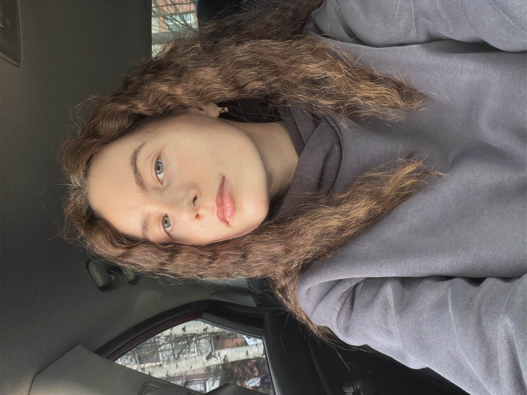 A person is positioned inside a vehicle, possibly a car, with a view to the outside through the window. The location is suggested to be Vsevolozhsk, Russia, based on the prompt. The person has curly brown hair and is wearing a gray hooded sweatshirt. The interior of the vehicle is visible, including the ceiling and window frame. The exterior view through the window includes buildings and trees.