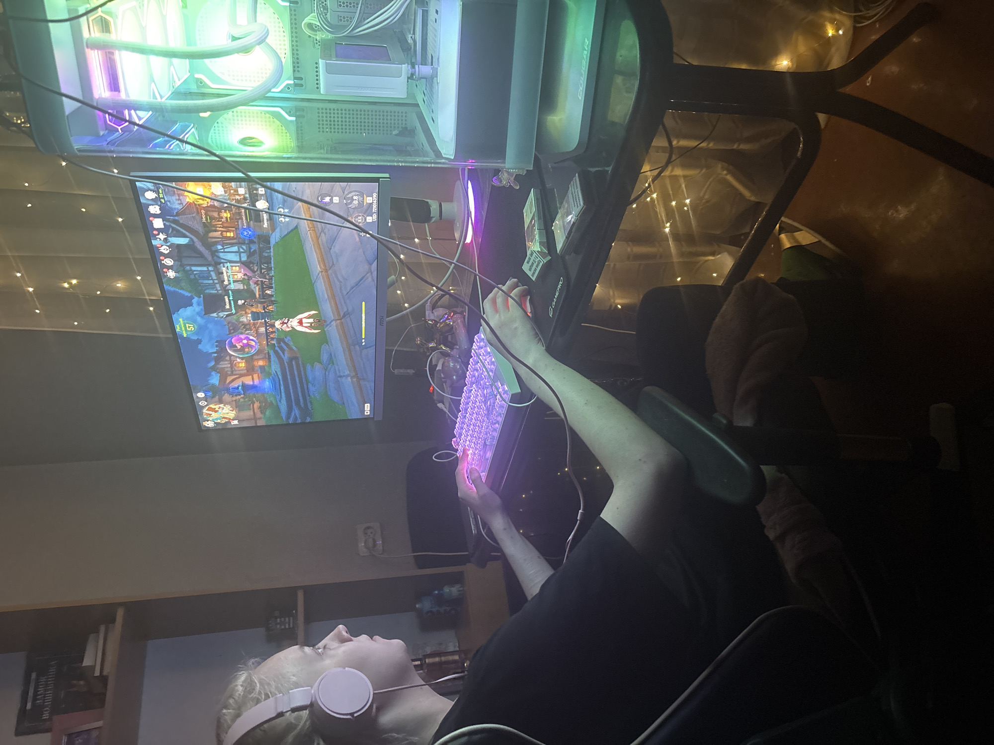 An individual, with light-colored hair pulled back and wearing pink headphones and a black t-shirt, is seated in a gaming chair, facing a computer monitor. The monitor displays a video game scene. Their right hand is positioned over a glowing pink keyboard, and their left hand is near a mouse. In front of the keyboard are two packs of cigarettes and a lighter. To the right of the monitor, a large, illuminated computer tower with green and purple lights is visible. The scene is set on a dark, glossy table with a black gaming mousepad. String lights are draped behind the monitor and around the table legs, casting a warm glow. The background shows a bookshelf with books and some decorative items. The overall environment appears to be a room, likely a home gaming setup, with low lighting.
