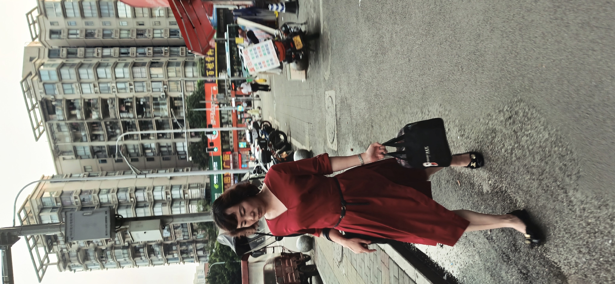 A woman in a red dress with a black belt and black shoes is walking on a paved street. She is carrying a black tote bag in her right hand. Behind her is a row of parked motorcycles and a street with buildings in the background. The location is the city of Jiulongpo District, China.