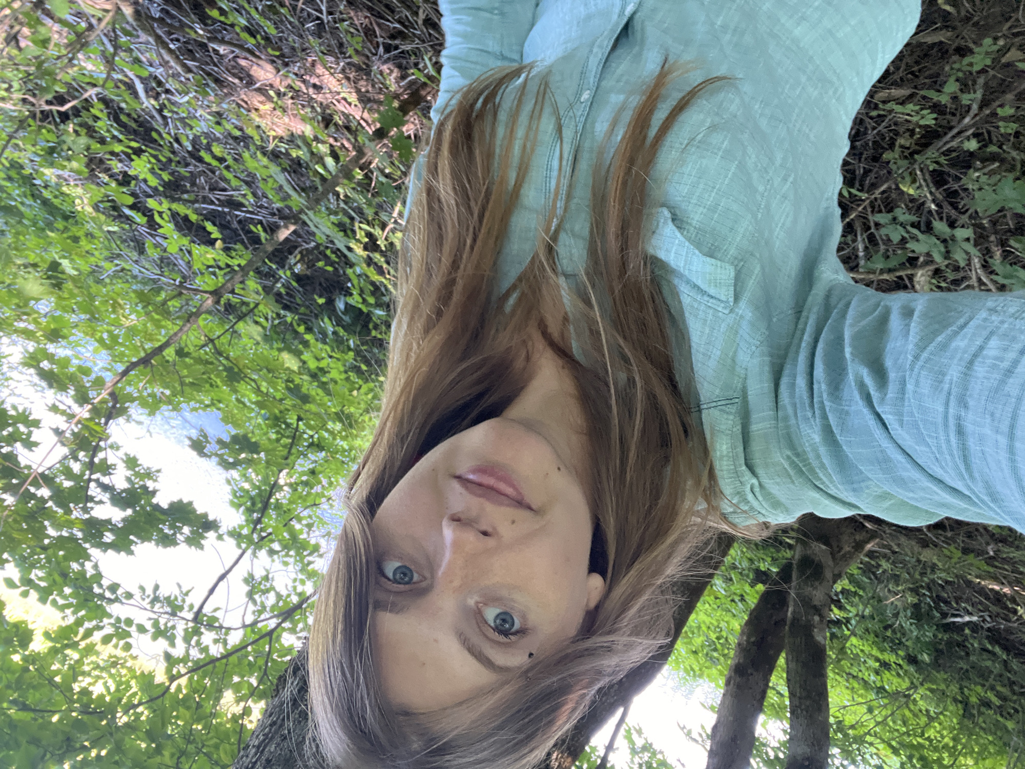 A woman is taking a selfie outdoors, with trees and foliage in the background. She has long, light brown hair and is wearing a light blue, button-up shirt. Her eyes are blue, and she has some freckles and a beauty mark above her left eye. The setting appears to be a wooded area, possibly a park or forest, with dense green leaves and branches. The ground is covered with fallen leaves and twigs. The image is taken from a slightly elevated angle. The specific location in Германавіцкі сельскі Савет, Belarus cannot be determined from the visual information in the image.