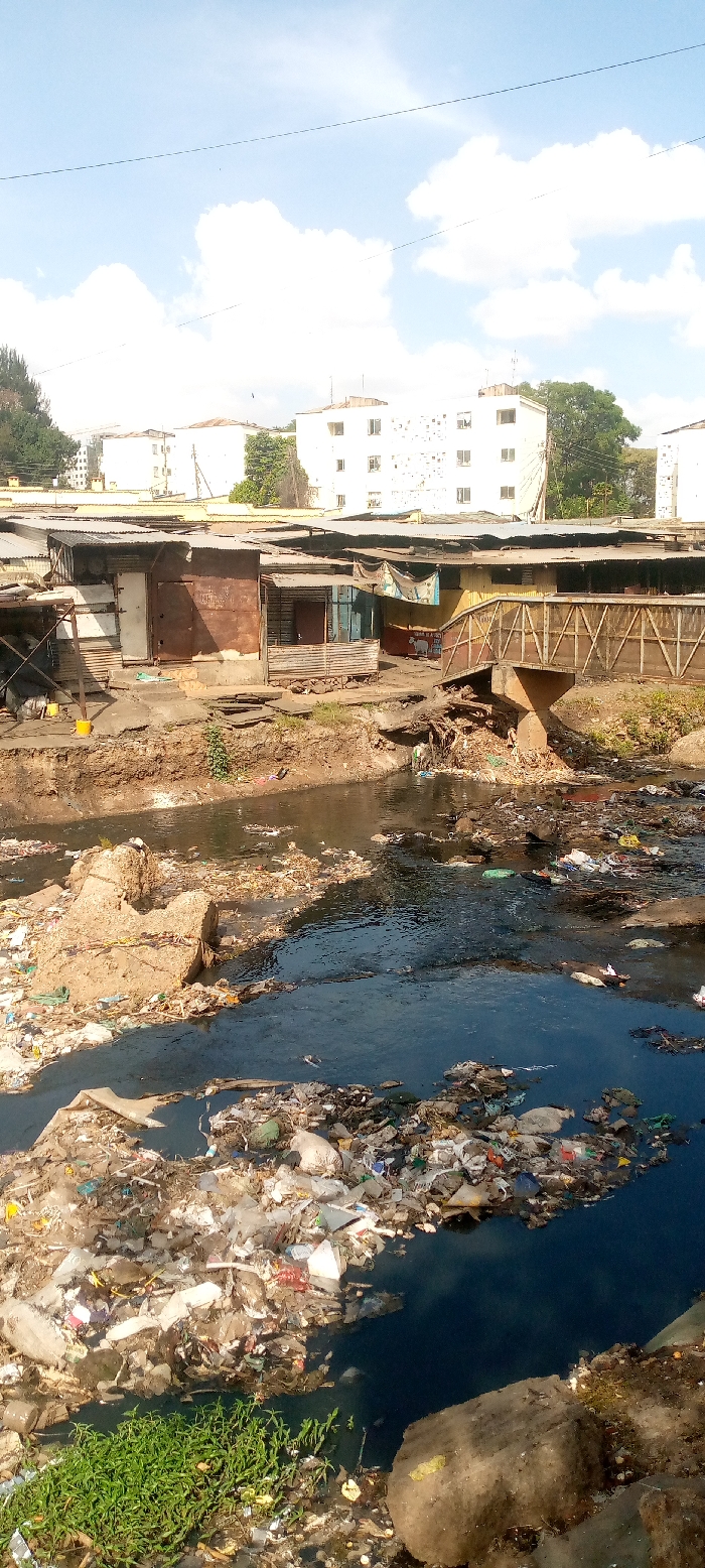 The image presents a wide view of a heavily polluted urban waterway, identifiable as a section of the Nairobi River in Nairobi, Kenya, under a partly cloudy blue sky. The foreground and midground feature the river, its dark water almost entirely obscured by a dense accumulation of solid waste, including plastic bottles, bags, and various debris, forming large floating islands of trash. The riverbanks are also heavily littered. On the far bank, a line of informal structures, likely shanties or market stalls, are constructed from corrugated metal sheets, wood, and other repurposed materials, topped with corrugated iron roofs. A functional, though rudimentary, metal truss bridge with wooden railings extends across the river, connecting to these structures. Beneath the bridge, near the heavily polluted waterline, a small, indistinct figure is visible. In the background, elevated above the informal settlement, several multi-story residential buildings with light-colored, weathered facades and multiple windows are present. No other direct human actions or interactions are clearly discernible within the frame. The overall environment indicates severe environmental degradation within a dense urban setting.