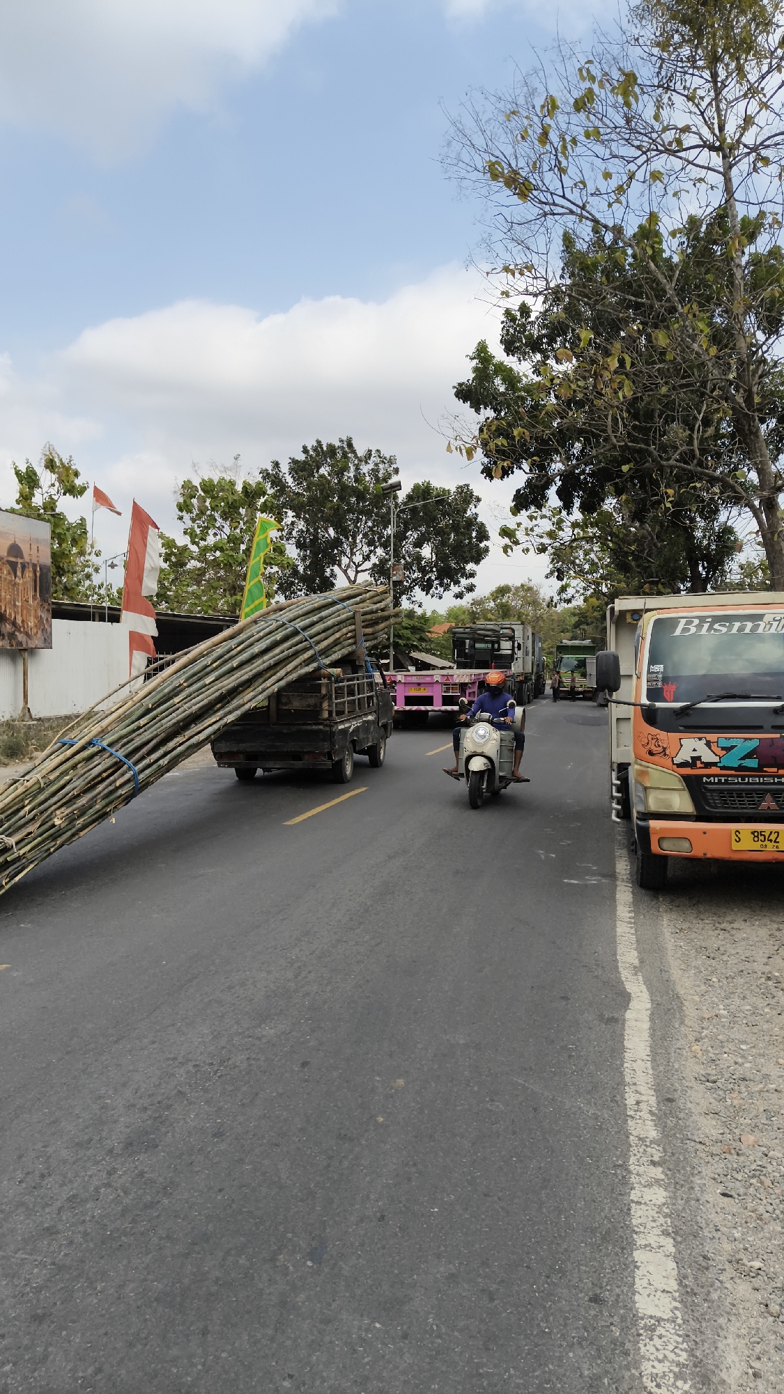 In Sambipondok, Indonesia, a street scene unfolds under a partly cloudy sky. A flatbed truck, heavily laden with a large bundle of bamboo poles secured by blue ropes, dominates the foreground. The truck is in motion on an asphalt road marked with yellow lines. A scooter rider, wearing a blue shirt and an orange helmet, is traveling behind the truck, also on the road. Several other trucks are visible in the background, including an orange Mitsubishi Fuso truck on the right side of the image, displaying the license plate "S 8542 SB". Trees line the right side of the road, and flags, including Indonesian flags, are visible in the distance. A billboard or large advertisement is partially visible on the left side of the frame. The environment suggests a sunny day with some scattered clouds.