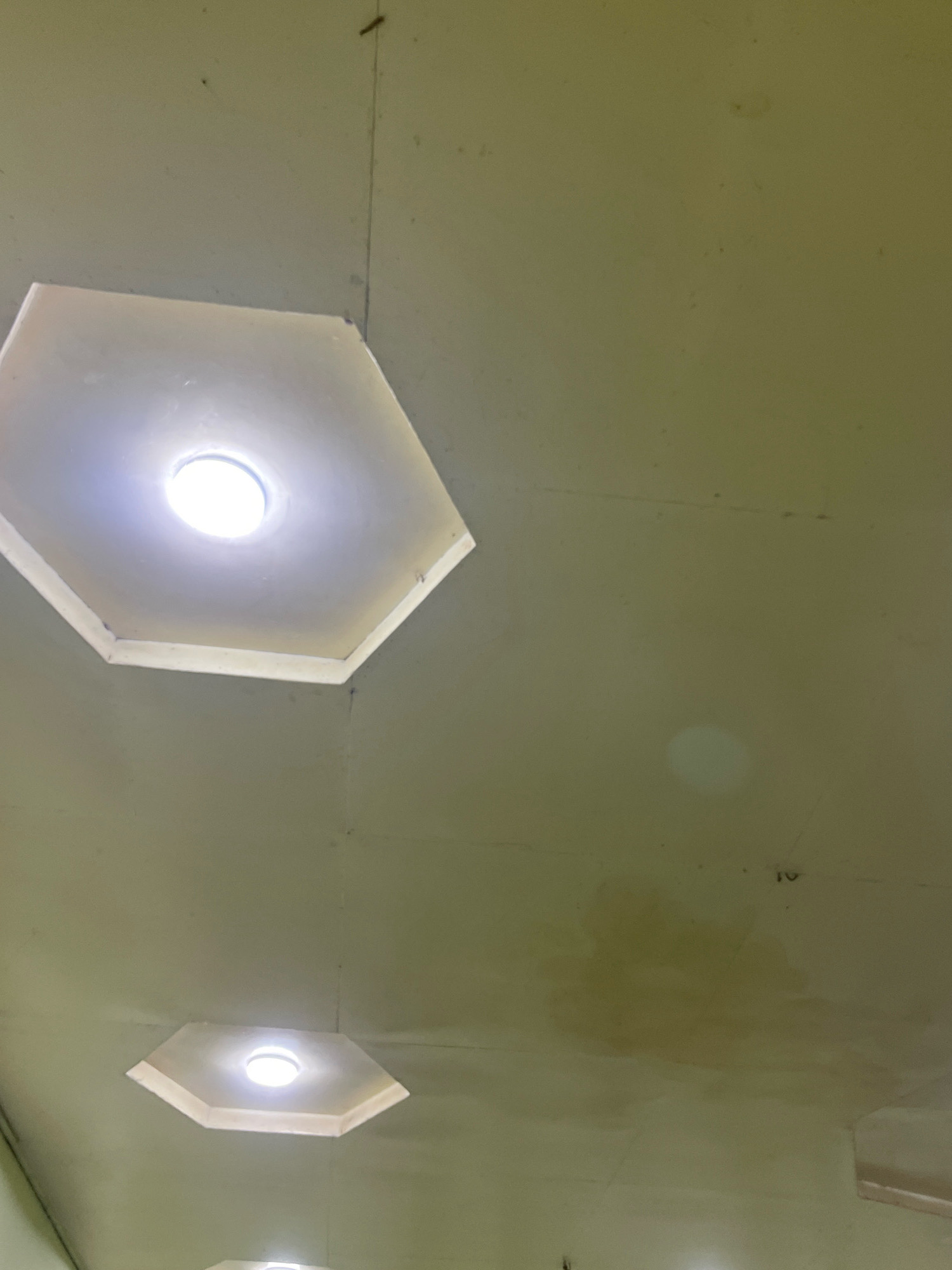 The image shows a ceiling with two hexagonal recessed lights. The ceiling is painted a light, muted green color and appears to be made of plaster. There are some minor imperfections and faint marks on the ceiling, including a water stain in the upper right quadrant. One light is in the upper left of the frame, and another is in the lower center. Both lights are illuminated, emitting a bright white light. The overall impression is of an indoor space, possibly a hallway or a room with a simple, functional design. No people or specific architectural features that would definitively place it in Пасариқ, Tajikistan are visible.