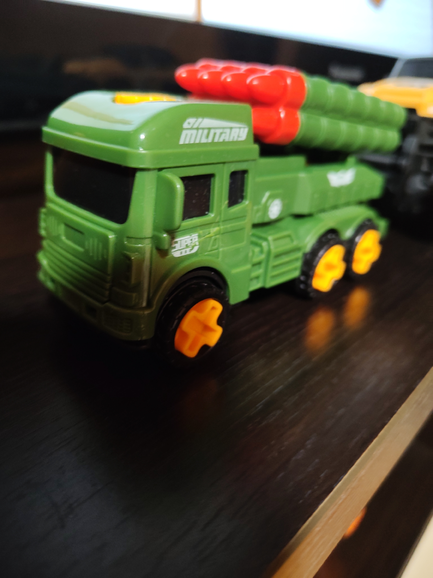 A green toy missile truck is shown from a low angle, placed on a dark wooden surface. The truck has red missiles on top and orange wheels. The word "MILITARY" is visible on the side of the truck. The background is out of focus and appears to be a screen or monitor, with a hint of another yellow toy vehicle to the right. The scene is indoors, with no visible indications of the city of Фестивальное Снт, Russia, or any people.
