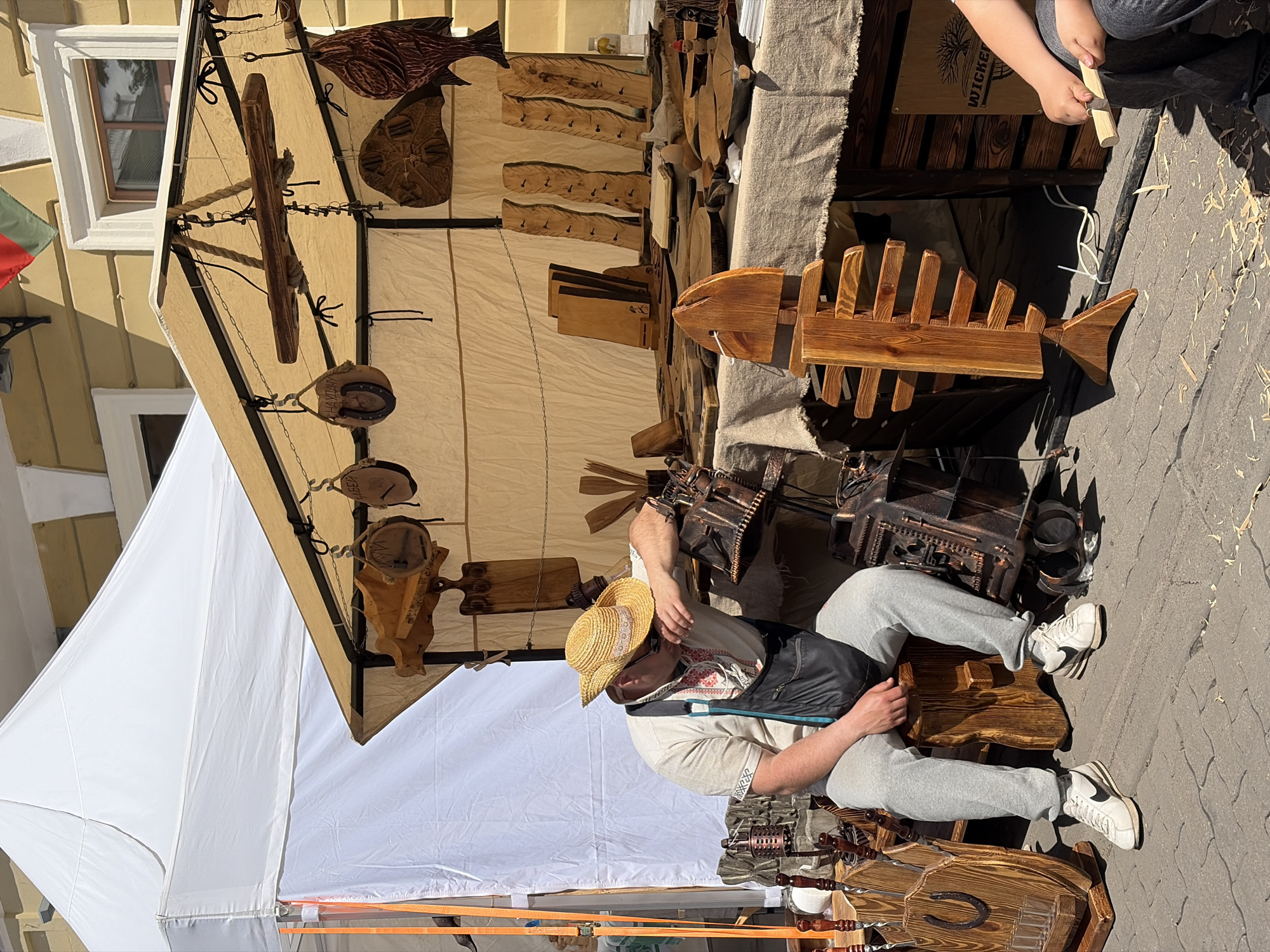 The image shows a man wearing a straw hat, light-colored shirt, dark apron, and gray sweatpants, seated on a wooden horse-shaped seat. He is positioned within a market stall covered by a large white and beige canopy. Hanging from the beige portion of the canopy are various wooden objects including fish, crosses, and other decorative items. Behind the man, on a table covered with gray fabric, are more wooden objects, including a decorative wooden fish. To the right of the table, a child is holding a wooden stick. The ground is paved with gray stones. Based on the style of crafts and the presence of a Bulgarian flag, the location could potentially be within a market or street fair in Minsk, Belarus.
