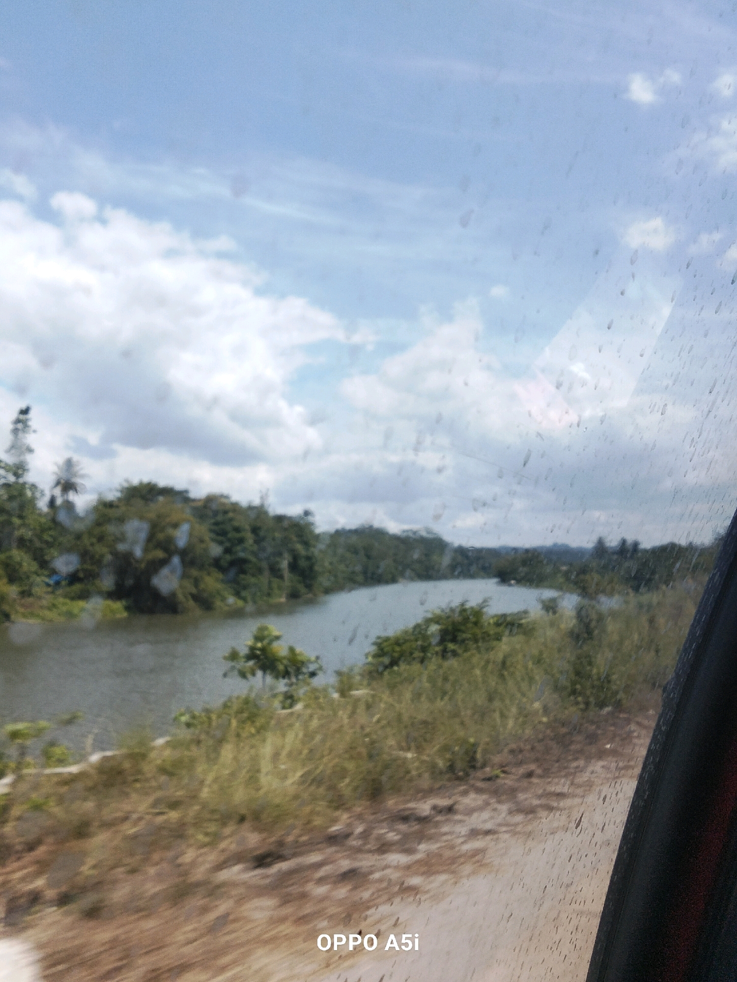 The image captures a scenic view of a river bordered by lush green trees and vegetation. The sky above is partly cloudy, with patches of blue visible. The foreground shows a dirt road or pathway, and a portion of a vehicle's interior, possibly a window with water droplets on it, is visible on the right side of the frame. The overall impression is of a natural landscape, possibly taken from a moving vehicle on a cloudy day. The timestamp "OPPO A5i" suggests the image was taken with a mobile phone of that model. There are no people or specific man-made structures clearly identifiable in the scene.