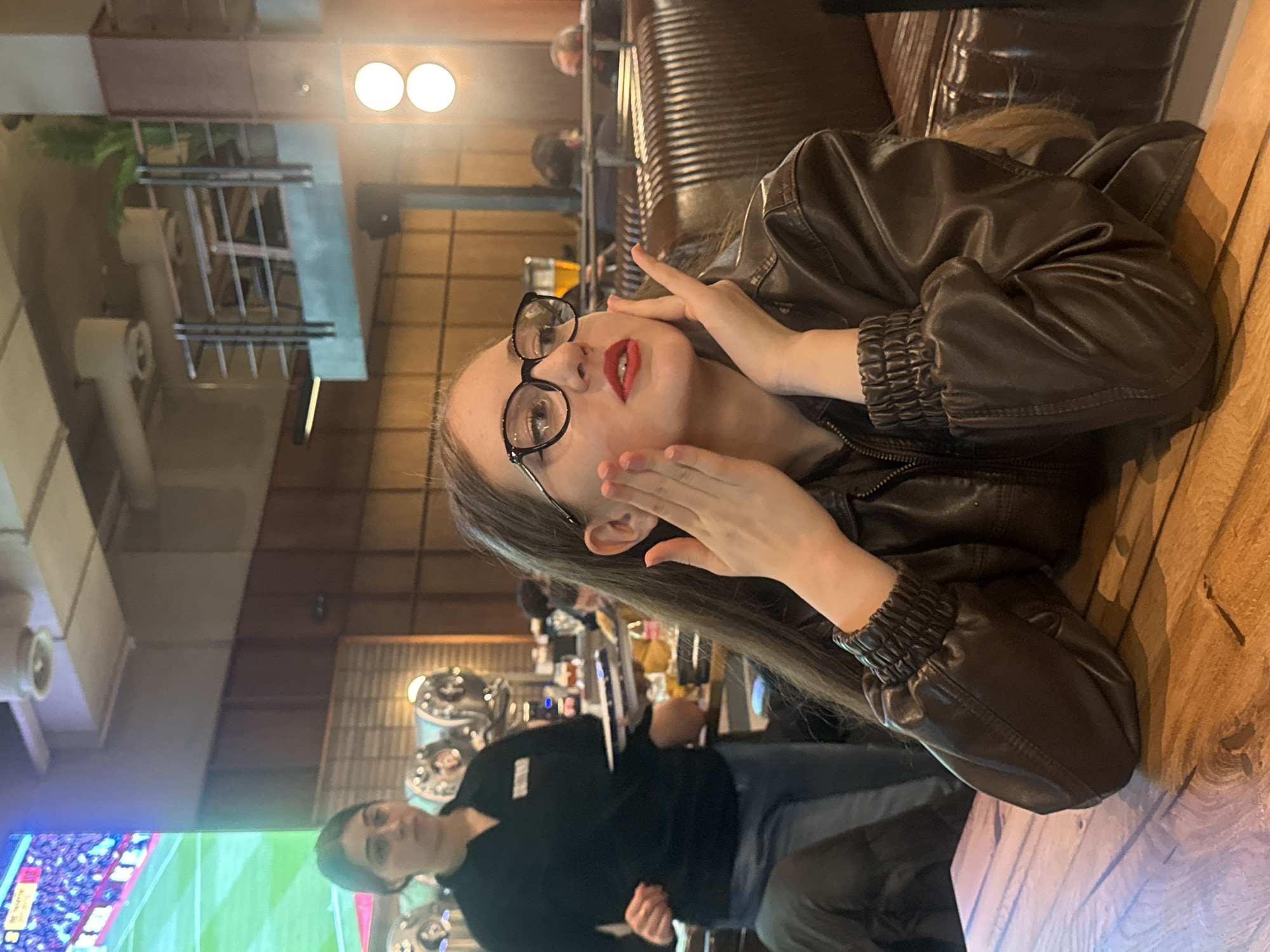 The image shows a person with long brown hair, wearing a brown leather jacket and black-rimmed glasses, with red lipstick, resting their hands on their face. They are at a table in a restaurant or bar. In the background, a server wearing a black shirt is visible, and a television screen displays a sports game. The setting appears to be a modern, dimly lit establishment with exposed wooden beams and industrial-style decor. The location is likely in Tbilisi, Georgia, but this cannot be confirmed from the image.