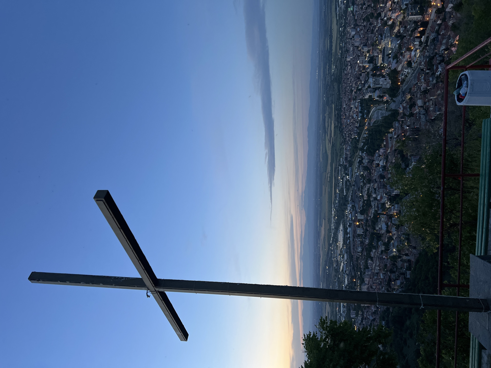 The image shows a view from a high point in Asenovgrad, Bulgaria, looking down over the city at dusk. In the foreground is a large metal cross. The sky is a gradient of blue, transitioning to orange near the horizon. A long, narrow cloud stretches vertically across the sky. Below the horizon, the city of Asenovgrad is visible, with buildings and lights dotting the landscape. In the lower right corner are trees and a red and green guard rail. A white trash receptacle is also partially visible.