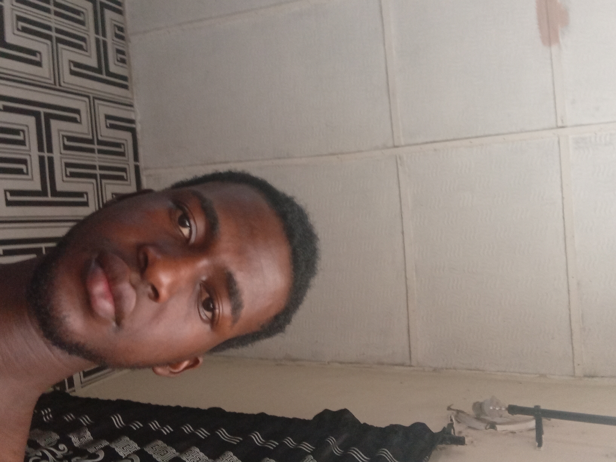 A close-up portrait shows a young Black man, seen from the upper chest up, posing for the camera. His head is tilted to the left, and he looks directly into the lens with a neutral to slightly pensive expression. He has short dark hair and a light beard or stubble.
The background is split between two distinct wall coverings. To the left, there's a patterned surface with a black and white geometric design, possibly wallpaper or a decorative panel. To the right and extending to the top, the wall is a plain, light-colored (possibly off-white) surface, divided into rectangular sections by horizontal and vertical lines, resembling large tiles or wall panels. A reddish-brown mark is visible on the upper right panel.
In the bottom foreground, a dark fabric object, which could be a blanket or clothing, features white wavy patterns and some geometric designs similar to those on the left wall. To the far bottom right, some indistinct objects, including what appears to be part of a metal pipe or bar, lie on a light-colored surface, possibly a floor or shelf. The lighting is even, suggesting indoor conditions, possibly natural light filtered from a window or artificial light. There is no visible text. The setting is an indoor space, located in Lokoja, Nigeria.