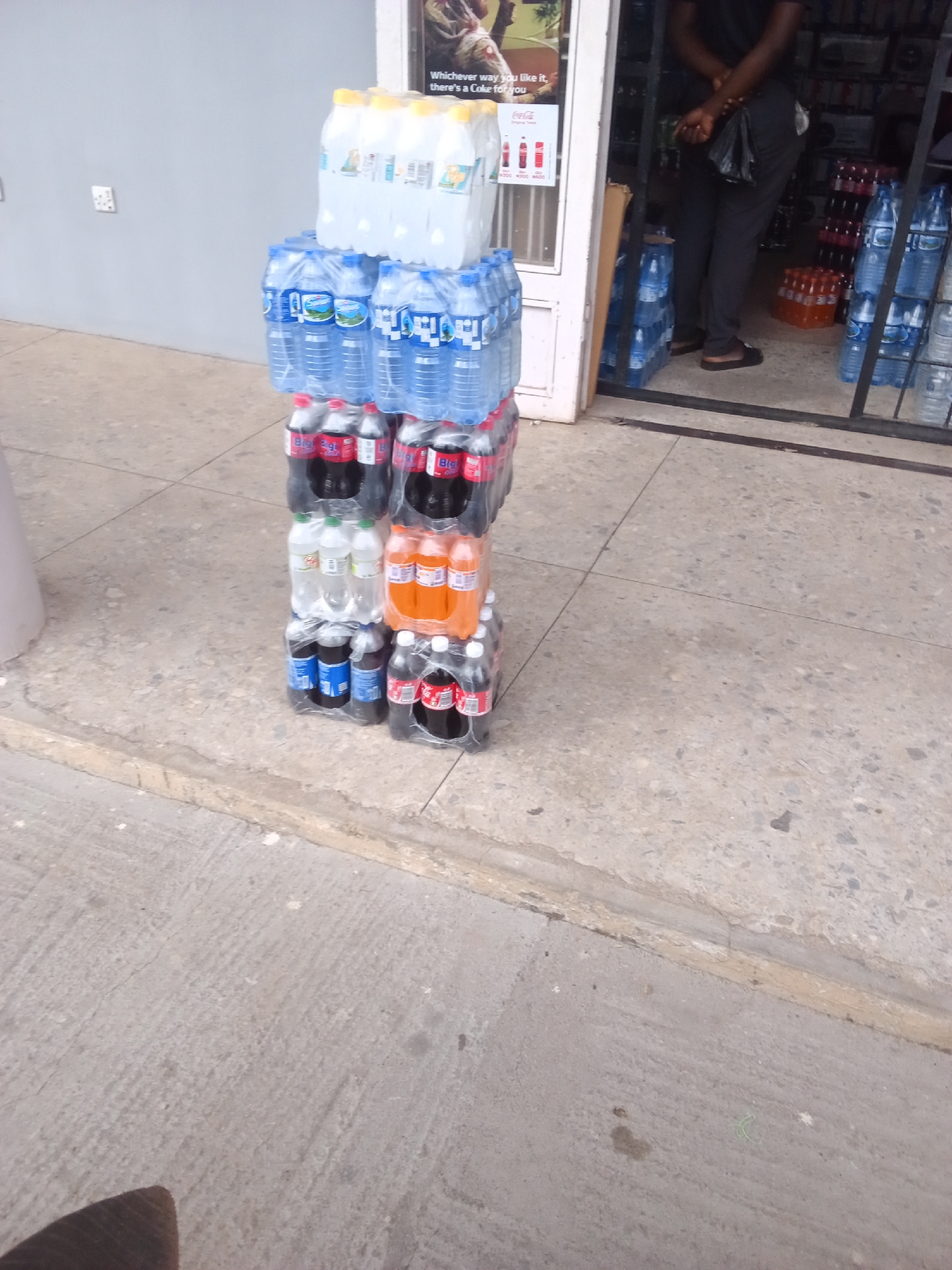 The image depicts a street scene in Jos, Nigeria, with a focus on stacked beverages outside a shop. In the foreground, several stacks of bottled drinks are arranged on the concrete pavement. The topmost stack appears to be bottled water, followed by stacks of soft drinks, including what seem to be Coca-Cola and other dark-colored beverages. A stack of orange-colored drinks is also visible. The bottles are primarily plastic and wrapped in cellophane.
In the background, a shop entrance is visible. A person is standing inside the doorway, with their arms crossed. The shop appears to be selling drinks, as suggested by the rows of bottles visible behind the person. A sign is partially visible above the doorway, with some text and an image, possibly an advertisement. The setting is an outdoor commercial area, with a textured concrete ground and a plain grey wall to the left. The lighting suggests it is daytime, but the weather is not discernible. The overall impression is of a typical retail environment in a Nigerian urban setting.