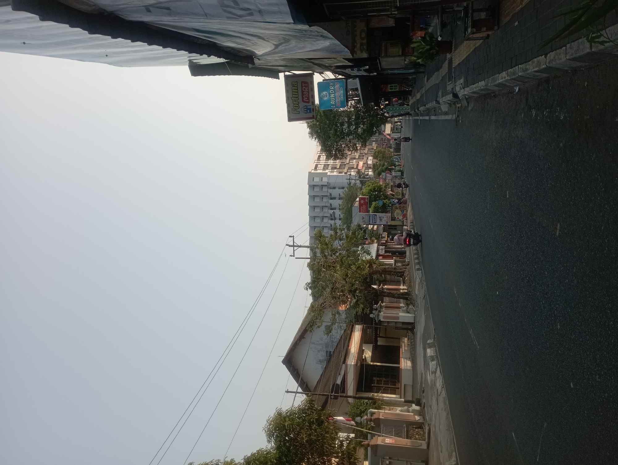 The image depicts a street scene in Yogyakarta, Indonesia, bathed in the soft light of what appears to be either early morning or late afternoon. The perspective is from the road, looking down a moderately busy street lined with buildings and trees.
On the left side of the street, a traditional-looking house with a white facade and a dark tiled roof stands out. It has a fenced entrance with stone pillars and an Indonesian flag planted near the gate. Further down the street on the left, more trees and buildings are visible, some with visible signage.
The right side of the street features a sidewalk and a row of shops. Signs for "Fortuna Mart" and "Joy Laundry Express" are prominently displayed, indicating commercial activity. The buildings on this side appear to be a mix of older structures and perhaps some newer developments. A large, light-colored building with a distinct geometric facade is visible in the background, suggesting modern architecture amidst the older streetscape.
Several people are present, primarily riding motorbikes on the street. One person is clearly visible on a motorbike in the foreground on the right side of the road, heading away from the viewer. Other motorbikes are seen further down the street, suggesting transportation is a primary mode of travel. Potted plants are placed along the sidewalk, adding a touch of greenery to the urban environment. The sky is clear and pale, indicating fair weather. The road surface is asphalt, marked with white lines. The overall impression is a typical urban street in Indonesia, with a blend of residential and commercial properties, and a common mode of transportation being motorbikes.
