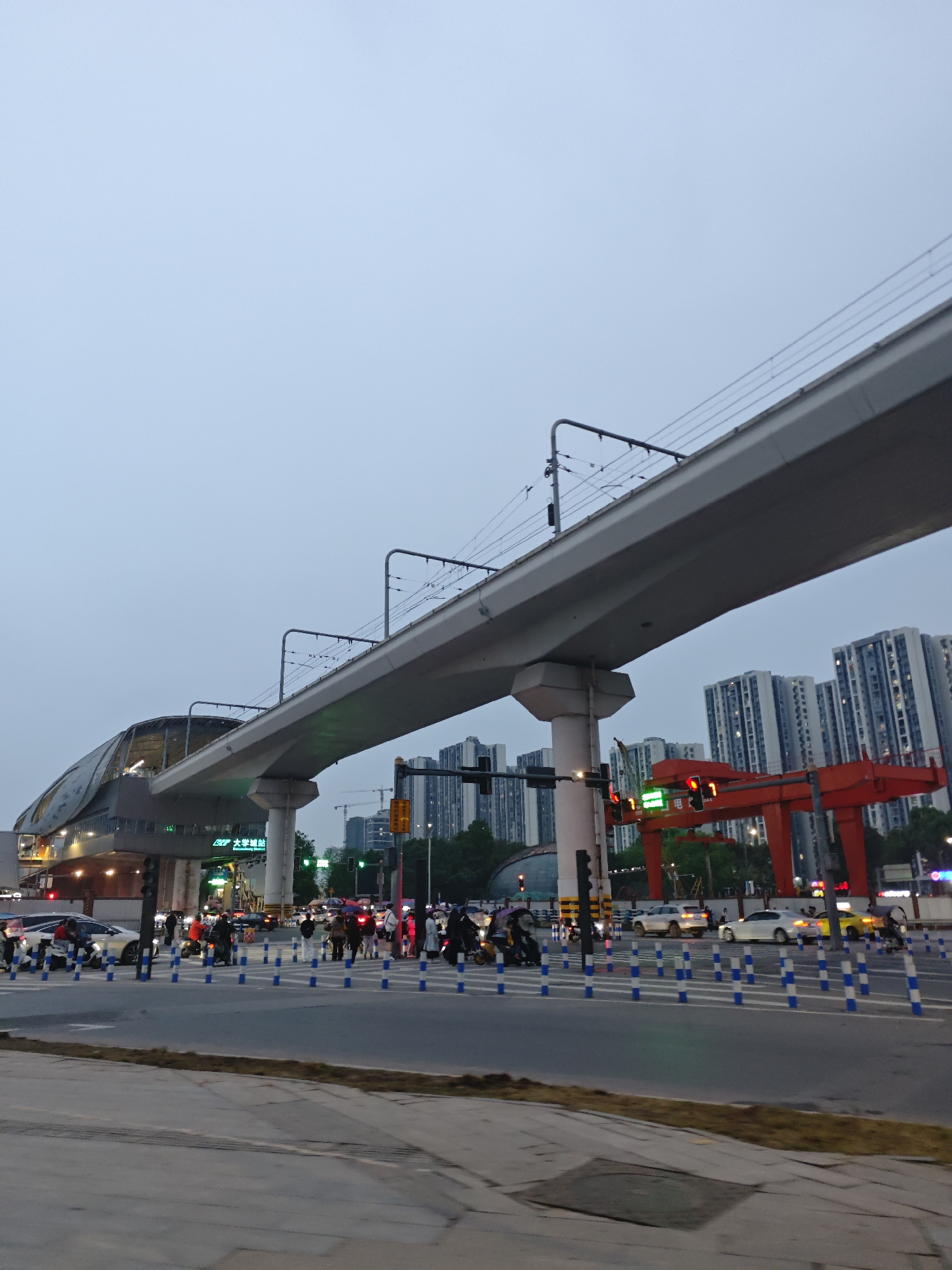 The image depicts an urban scene in Chongqing High-tech Zone, China. An elevated monorail structure dominates the view, with a station visible to the left. Below the monorail, a busy intersection is filled with pedestrians and vehicles. The intersection features blue and white traffic bollards. Buildings, including residential high-rises, form the backdrop, and the sky is overcast. A large, red gantry structure is situated near the intersection. The overall atmosphere suggests dusk or early evening.