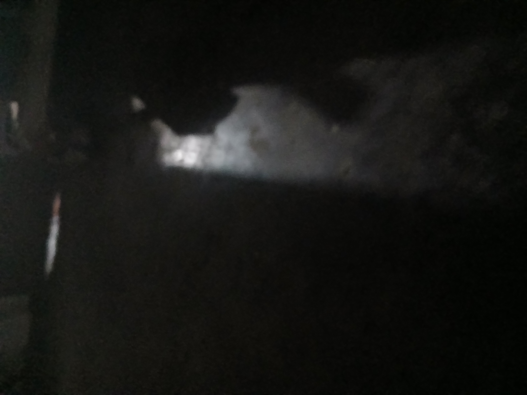A dimly lit, indoor scene in Kademangan Kulon, Indonesia, shows a silhouette of a person in the right half of the frame, with their back to the viewer. The person appears to be wearing dark clothing and is standing in what looks like a shaded area. A faint light source illuminates the floor in front of them, casting a shadow that suggests movement. The background is dark and indistinct, with no discernible architectural details or other objects. The overall impression is one of low light conditions, potentially at night or in a poorly lit interior space.