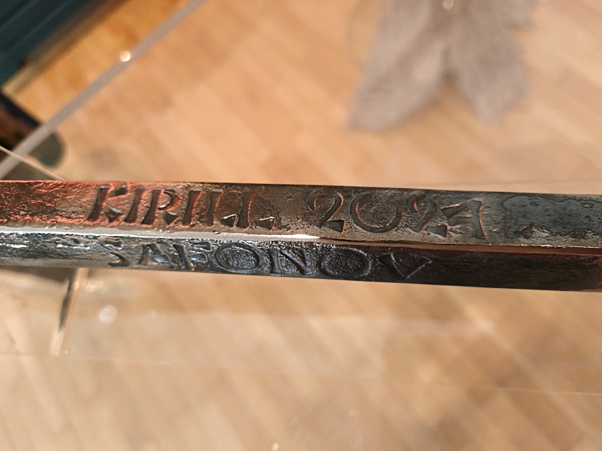 The image shows a close-up view of an engraved metal object, possibly a sword or a decorative blade. The engraving appears to read "KIRILL. 2021" on the upper line and "SAFONOV" on the lower line. The metal is dark and appears aged or weathered. The background consists of a wooden surface, likely a table or shelf, with a soft focus and warm lighting. The object is positioned horizontally across the frame, with a clear reflection visible on its surface, suggesting it is placed on a smooth, possibly glass, surface. No specific location in Tallinn, Estonia, can be determined from the visual information provided.