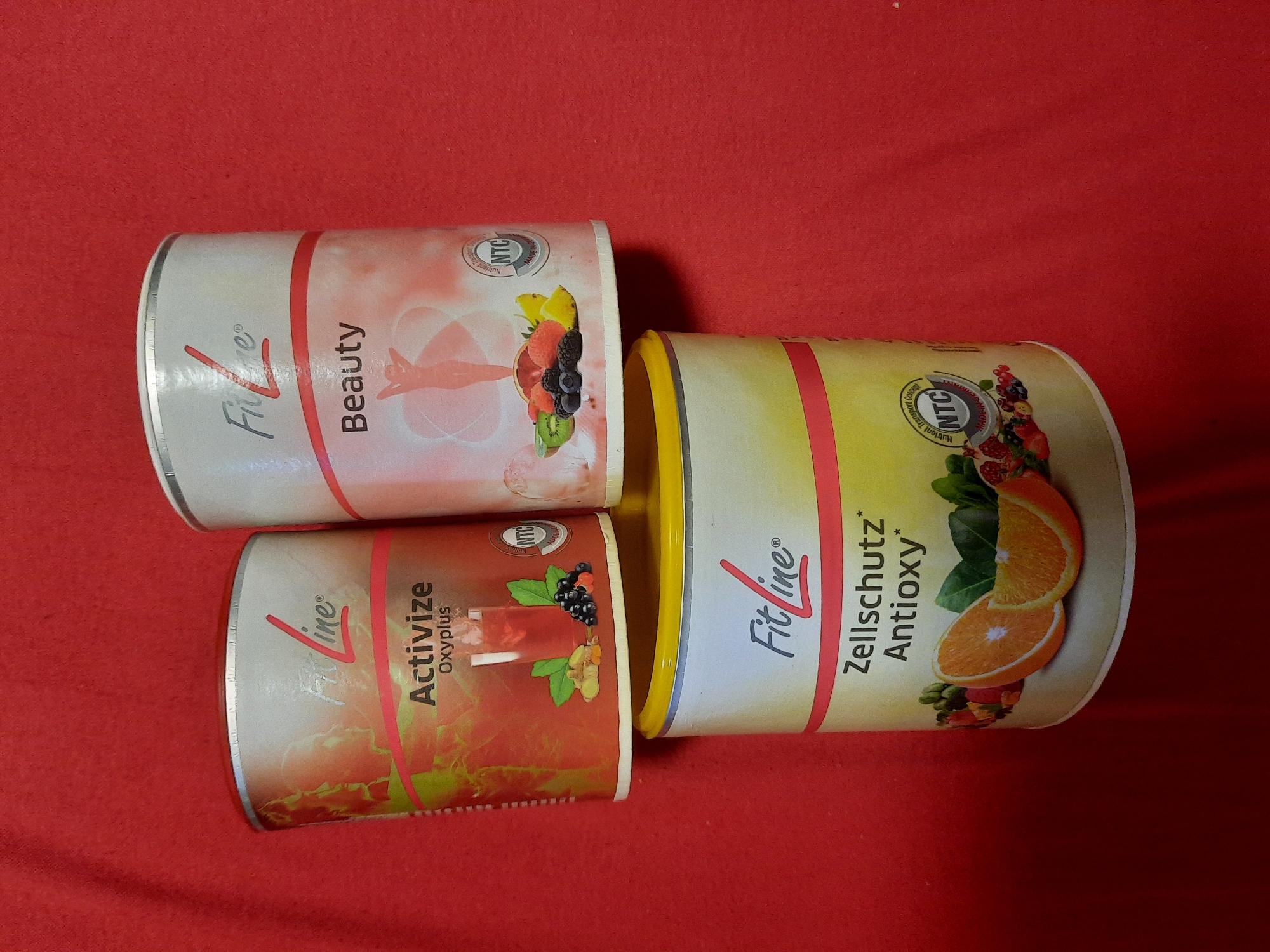 In a top-down shot, three cylindrical containers of FitLine products are arranged on a red textured surface. The top two containers are stacked side-by-side, with the bottom container directly beneath them, slightly offset to the left. All three containers feature prominent FitLine branding and product names. The top-left container is labeled "Activize Oxyplus" and displays an image of a drink with ice, ginger, and berries. The top-right container is labeled "Beauty" and features an illustration of a person's silhouette surrounded by swirling lines and an assortment of fruits including kiwi, grapefruit, and berries. A circular "NTC" logo is visible on both top containers. The bottom container is larger and appears to be a different product line, labeled "Zellschutz Antioxsy" and showcases images of oranges and various berries. No people or explicit location details of Rosenheim, Germany are visible in the image.