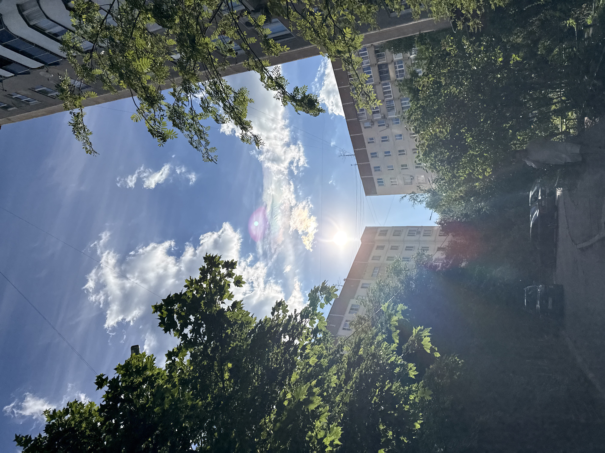 The image shows a view looking up towards the sky between two apartment buildings, with trees on either side framing the scene. The sun is visible in the sky, slightly off-center, creating a lens flare. There are white, puffy clouds against a blue sky, with thin, white contrails visible. The buildings appear to be multi-story residential structures with visible windows and balconies. Lush green trees and foliage are prominent in the foreground and mid-ground, with some sunlight filtering through the leaves. In the lower part of the image, a paved walkway or road is visible, with a few parked cars and a person walking. The overall impression is a residential urban environment on a sunny day.