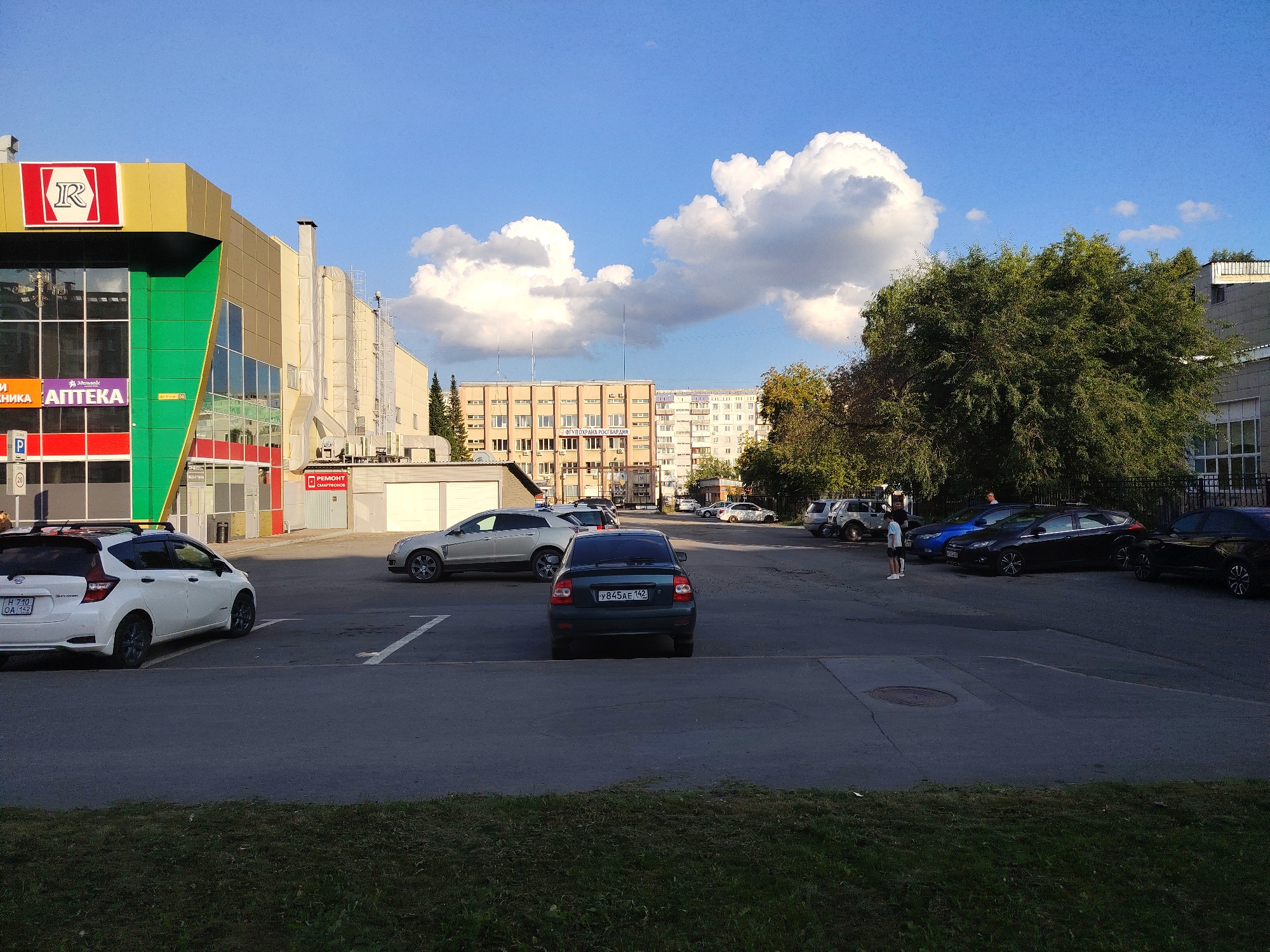 This is a ground-level, eye-level, full shot of a parking lot in front of a commercial building. The building is modern with a yellow and green facade and large windows. There are several cars parked in the lot, including a white Nissan Note on the left, a dark sedan in the middle, and a black sedan on the right. A person is standing in the parking lot near a blue car. The sky is blue with some white clouds. The image appears to be taken in Kemerovo, Russia, based on the license plate of the white car, which reads "H 710 OA 142". The license plate format is consistent with Russian license plates.