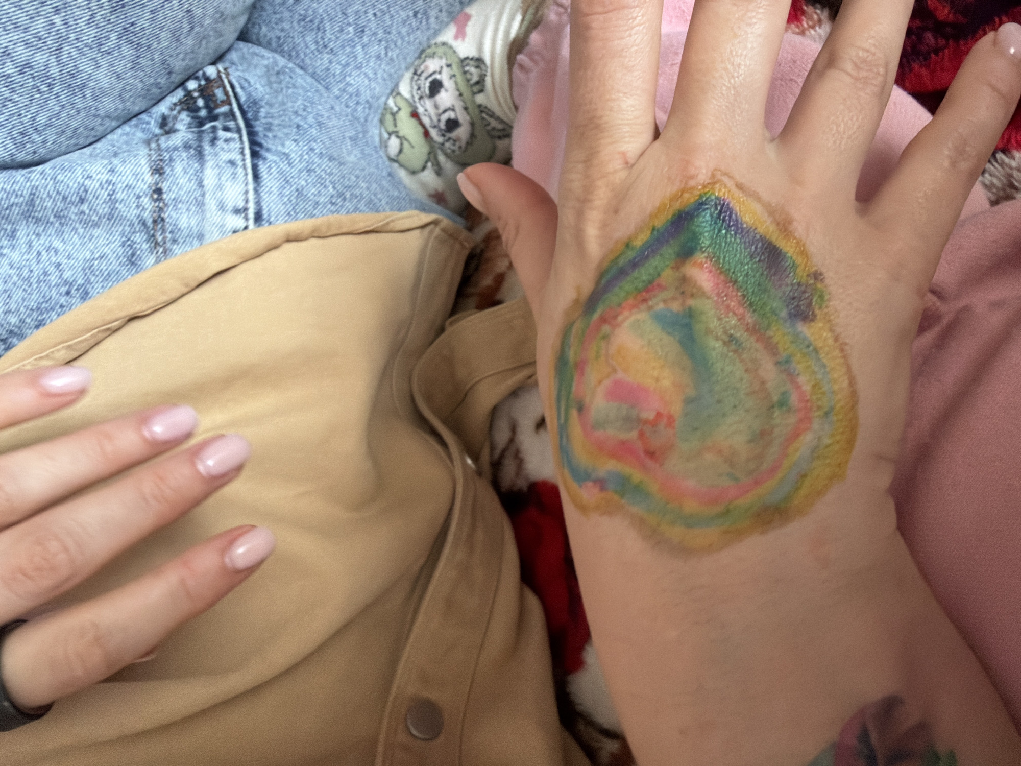 The image shows a close-up of a person's hand with a colorful, circular design on it. The design appears to be a temporary tattoo or body paint, featuring swirling patterns of yellow, green, blue, pink, and red. The hand is resting on a pink fabric. In the background, parts of blue denim clothing and a tan jacket are visible. The person's fingernails are painted a light pink color. There are no discernible elements in the image that would indicate a specific location within Valozhyn, Belarus.
