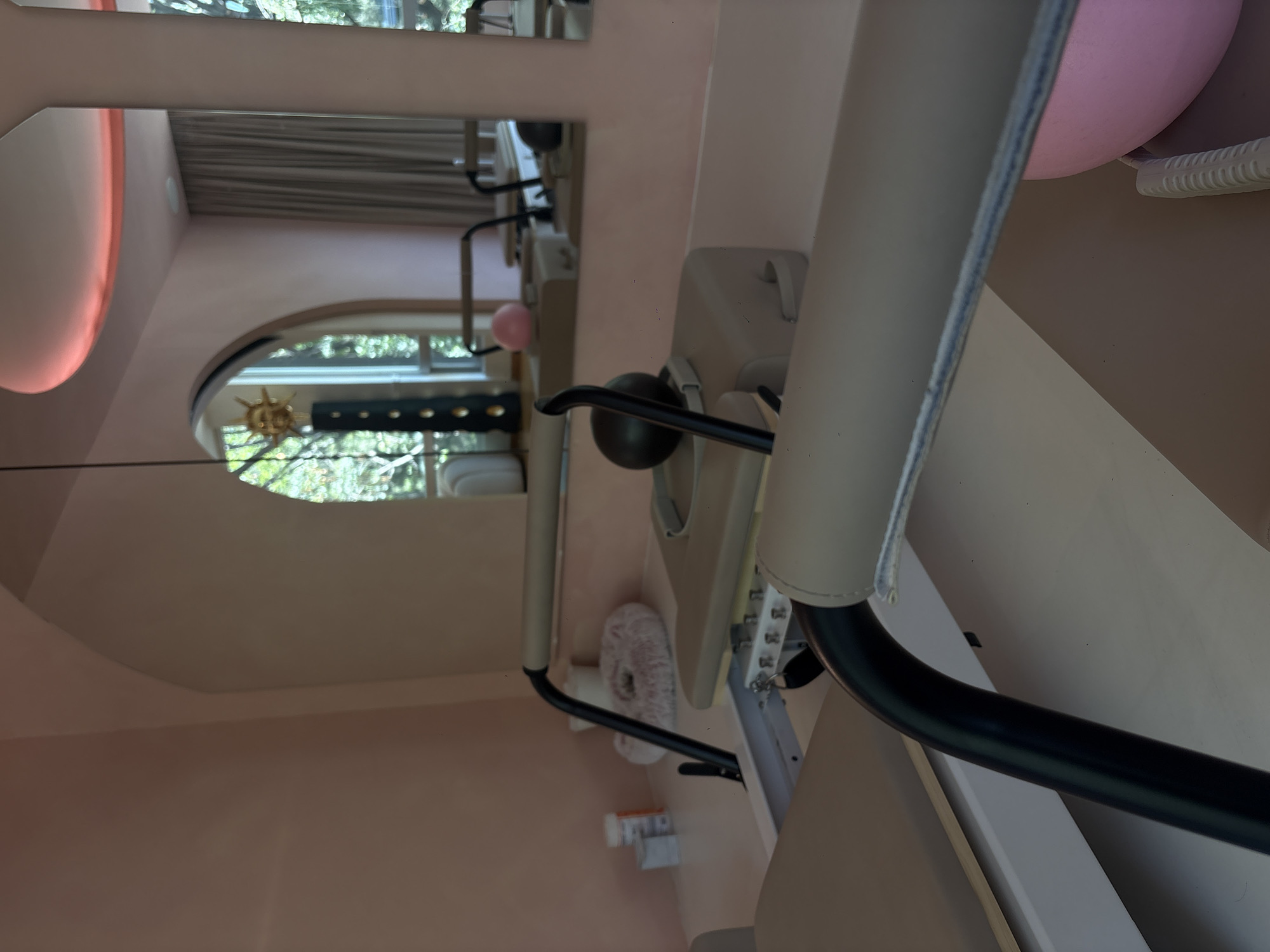 The image shows a pilates reformer machine in a room with pink walls. The machine is white and gray, with black handles and a pink balloon attached. The room appears to have a mirror on the wall that reflects a window with trees outside. The location is potentially Mexico City, Mexico.