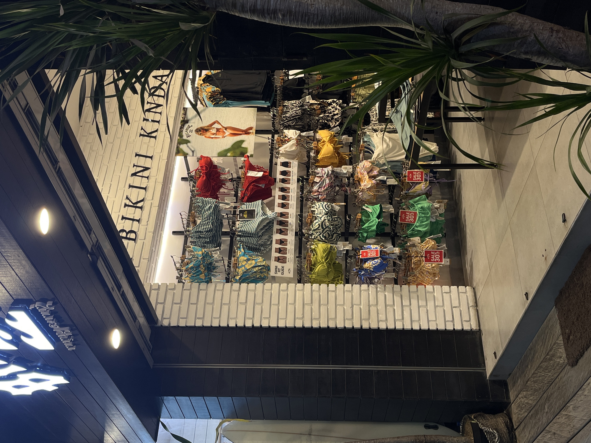 The image shows the exterior of a retail store in North Kuta, Indonesia, specializing in swimwear. The store's facade is predominantly black wood paneling on the left and white brick on the right, with a large opening displaying merchandise. Above the opening, the store's name, "BIKINI KINDA," is emblazoned in black lettering on the white brick wall.
Inside the store, numerous racks of bikinis and swimwear are visible. The bikinis are displayed in a variety of colors and patterns, with some featuring "SALE 30%" tags. A large advertisement poster of a woman in a white bikini is also displayed on the back wall.
The entrance to the store is at street level, with a slight step leading up to the interior. In front of the store, on the left, is a surfboard leaning against the wall and a woven basket. A large potted plant with long, green leaves stands to the right of the store entrance, partially obscuring the view of the interior. The ground in front of the store is paved with large, light-colored tiles, with a dark wooden plank walkway leading away from the entrance. The lighting suggests it is evening or nighttime, with artificial lights illuminating the store's interior and a portion of the exterior signage.