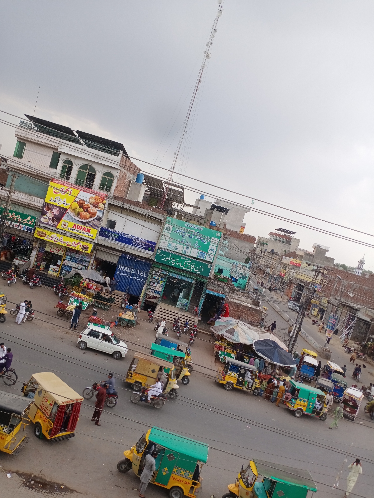 This image captures a bustling street scene in Farooqabad, Pakistan, under an overcast sky. The street is lined with multi-story buildings, many housing shops on their ground floors. Overhead, a dense network of electrical wires and communication cables crisscrosses the view.
Numerous people are visible, engaged in various daily activities. Pedestrians walk along the street and sidewalks, while others are seen interacting near market stalls. Many individuals are dressed in traditional South Asian attire. The street is filled with vehicles, predominantly auto-rickshaws (some marked "TEZRAFTAR"), motorcycles, and a white car. Several pushcarts laden with fresh produce like fruits and vegetables are positioned along the roadside, suggesting an active marketplace.
Key businesses visible include "AWAN SWEETS & BAKERS," prominently displayed on a large yellow and red sign, and "IRAC & TEL COMMUNICATIONS" on a blue sign. Another green sign identifies "دین فارمیسی سپر سٹور" (Din Pharmacy Super Store). Solar panels are visible on the roof of one building, and a mosque minaret can be seen in the distance, providing a characteristic architectural element of the region. The overall scene depicts a vibrant and active commercial area during what appears to be daytime, characterized by movement and commerce.