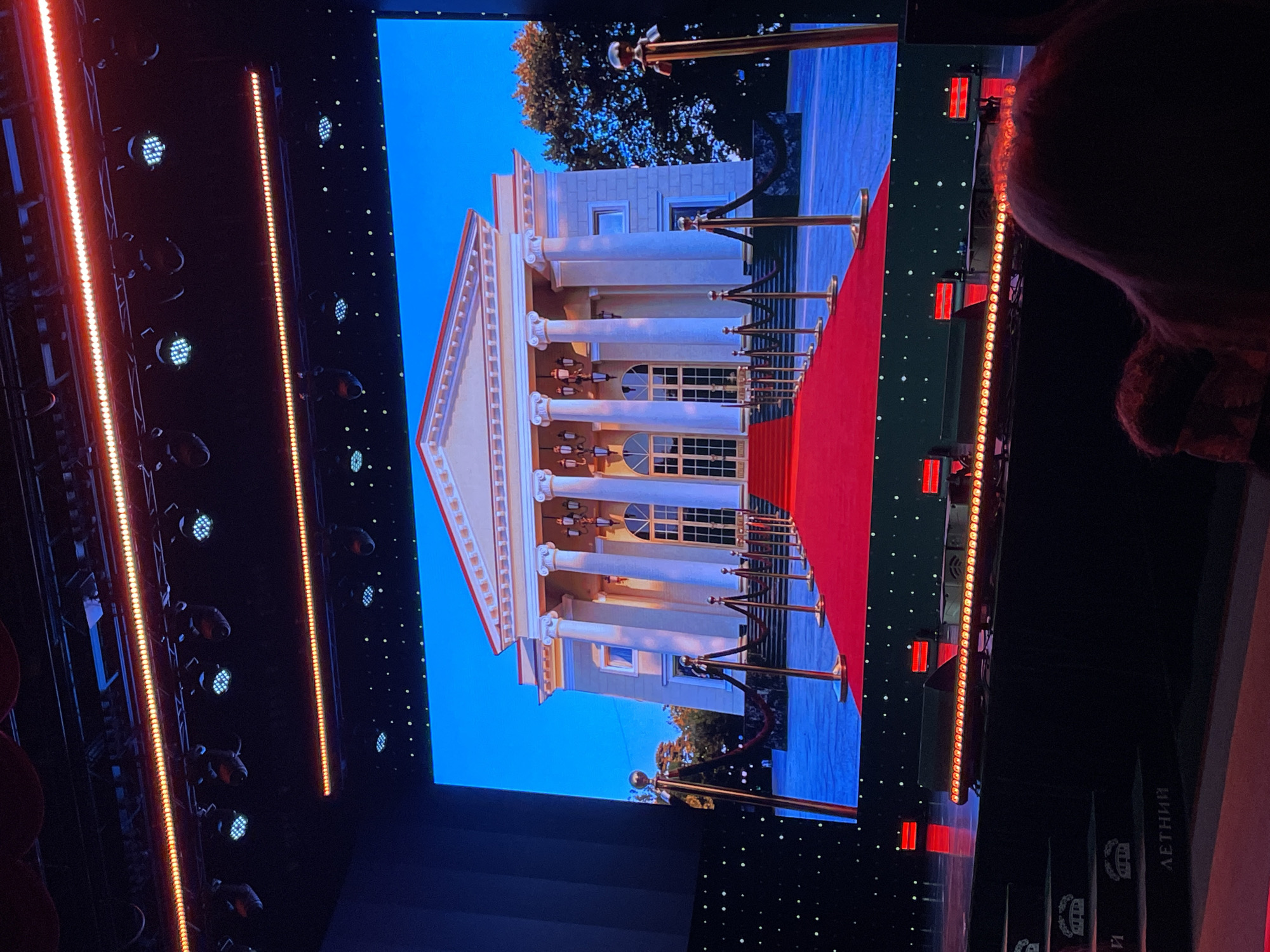 The image depicts a scene on a stage, likely within a venue in Sochi, Russia, given the context of past events. The primary focus is a large LED screen displaying a virtual representation of a grand building with prominent white columns, a red carpet leading up to its entrance, and ornate gold stanchions and velvet ropes. The building appears to be a neoclassical structure, possibly a palace or a theater. In the foreground of the stage, actual stanchions and velvet ropes are set up, mirroring the virtual display. There are also steps leading down from the stage, some of which have text labels visible, potentially indicating event areas or seating sections. The upper part of the image shows stage lighting equipment, including rows of orange and blue lights, and spotlights. In the very bottom right corner, the back of a person's head and shoulders are visible, indicating that the image was taken from an audience perspective. The overall impression is of a sophisticated event setup, possibly a red carpet premiere or awards ceremony.