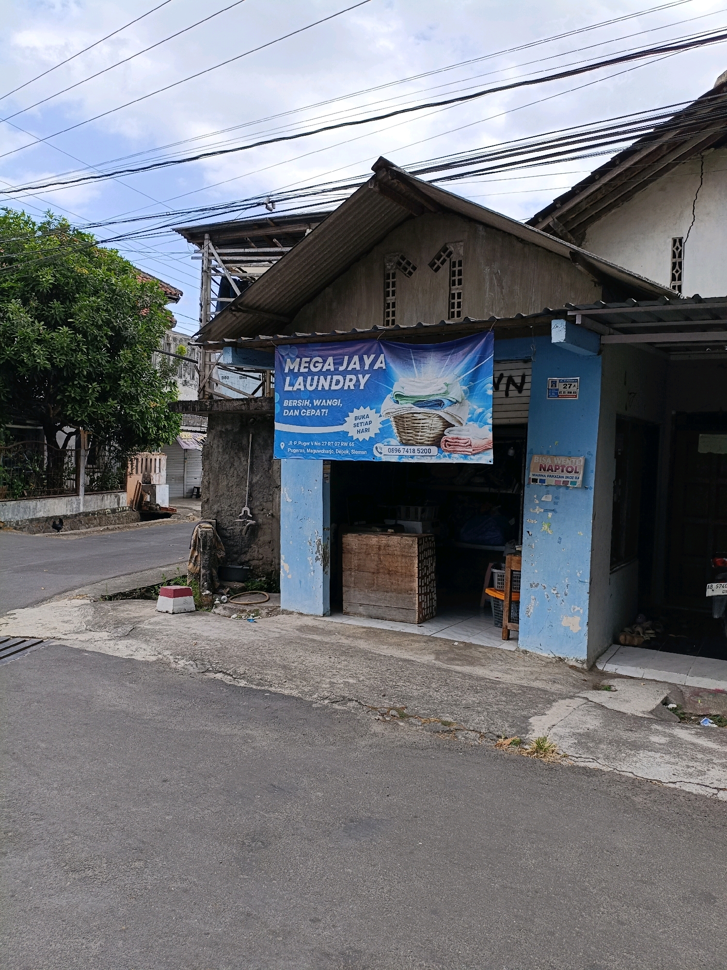 The image shows a street view in Maguwoharjo, Indonesia, with a laundry service storefront. A blue banner advertising "MEGA JAYA LAUNDRY" is prominently displayed on the building's facade. The laundry offers services described as "BERSIN, WANGI, DAN CEPAT!" (Clean, Fragrant, and Fast!). Below this, the address is listed as "Jl. P. Puger V No. 27 RT. 07 RW. 65, Pugeran, Maguwoharjo, Depok, Sleman." A phone number, "0896 7418 5200," is also visible.
The building itself appears weathered, with peeling blue paint on a pillar. Inside the open storefront, laundry baskets and what appear to be folded clothes are visible. To the right of the laundry entrance, there's a smaller sign that says "BISA NEGNTOL WARNA PAKAIAN JADI BA." (Can be blended, clothing colors become...). A motorbike with a license plate starting with "AB" is parked near the entrance.
The surrounding environment includes a paved road in the foreground, a sidewalk area with some vegetation and debris, and a large tree with dense foliage to the left. Power lines crisscross the sky above the buildings. The architecture suggests a typical Indonesian urban setting. The time of day appears to be daytime, given the natural light. There are no people clearly visible in the image.