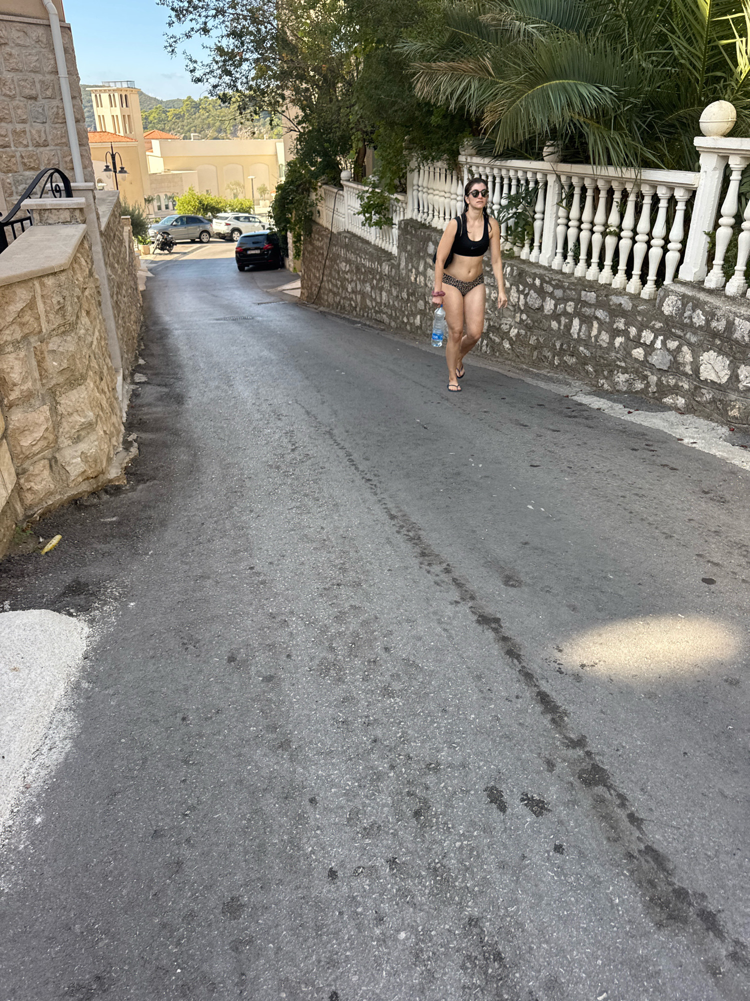 A woman in a black bikini top and leopard-print bottoms walks down a paved street in Boreti, Montenegro. She is holding a clear plastic bottle of water. The street slopes downhill and is lined on the left by a stone wall and on the right by a white ornate railing and a stone wall. Palm trees and other foliage are visible in the background. Several cars are parked on the side of the street further down. The sky is blue with some light clouds.