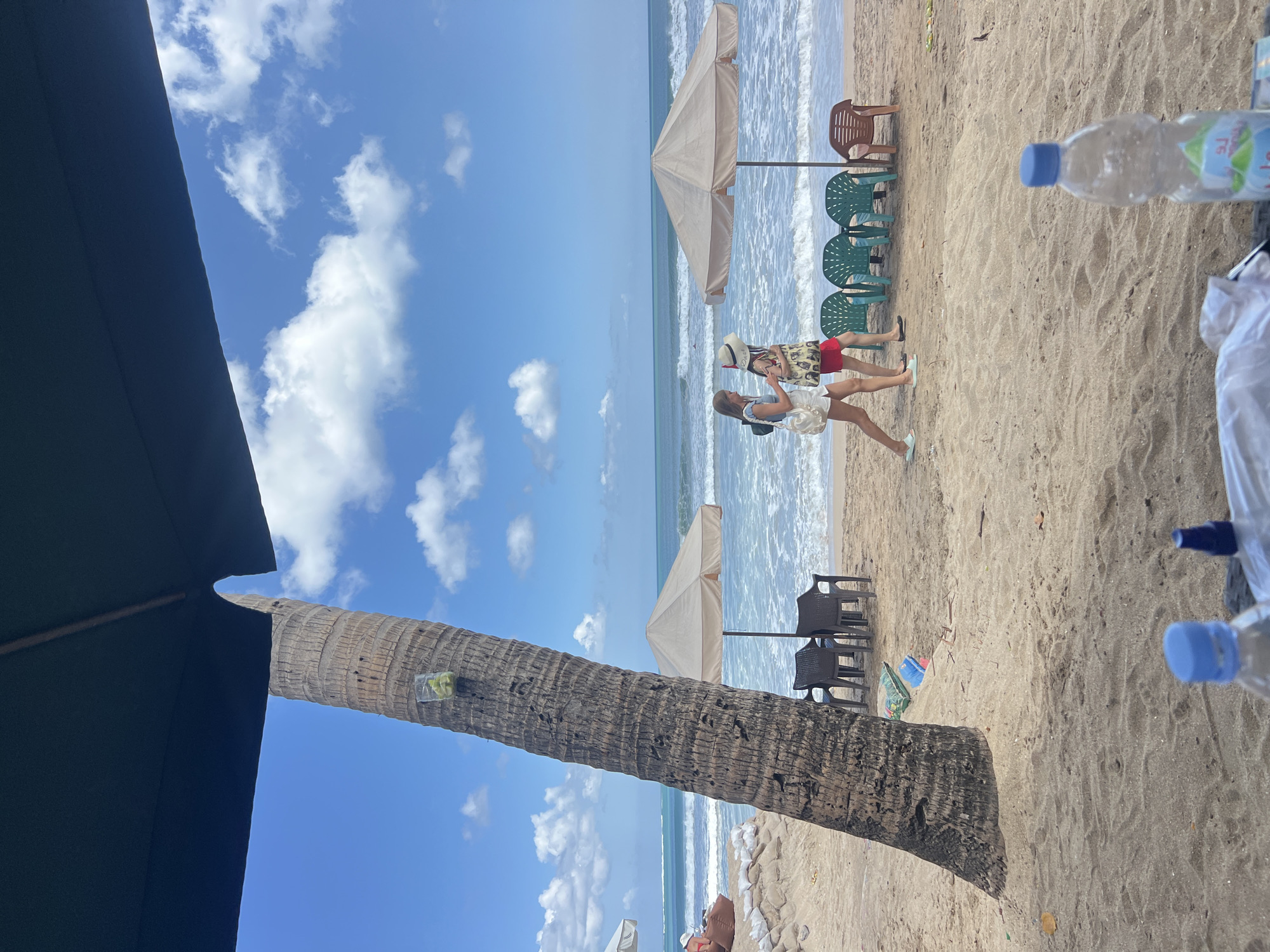 A beach scene in Kuta, Indonesia, shows two women walking on the sand near the ocean. A large palm tree stands in the foreground, with a dark umbrella casting a shadow over the scene. Beach umbrellas and chairs are set up on the sand, and the ocean waves are visible in the background. The sky is blue with scattered clouds. There are two bottles of water on the sand in the foreground.