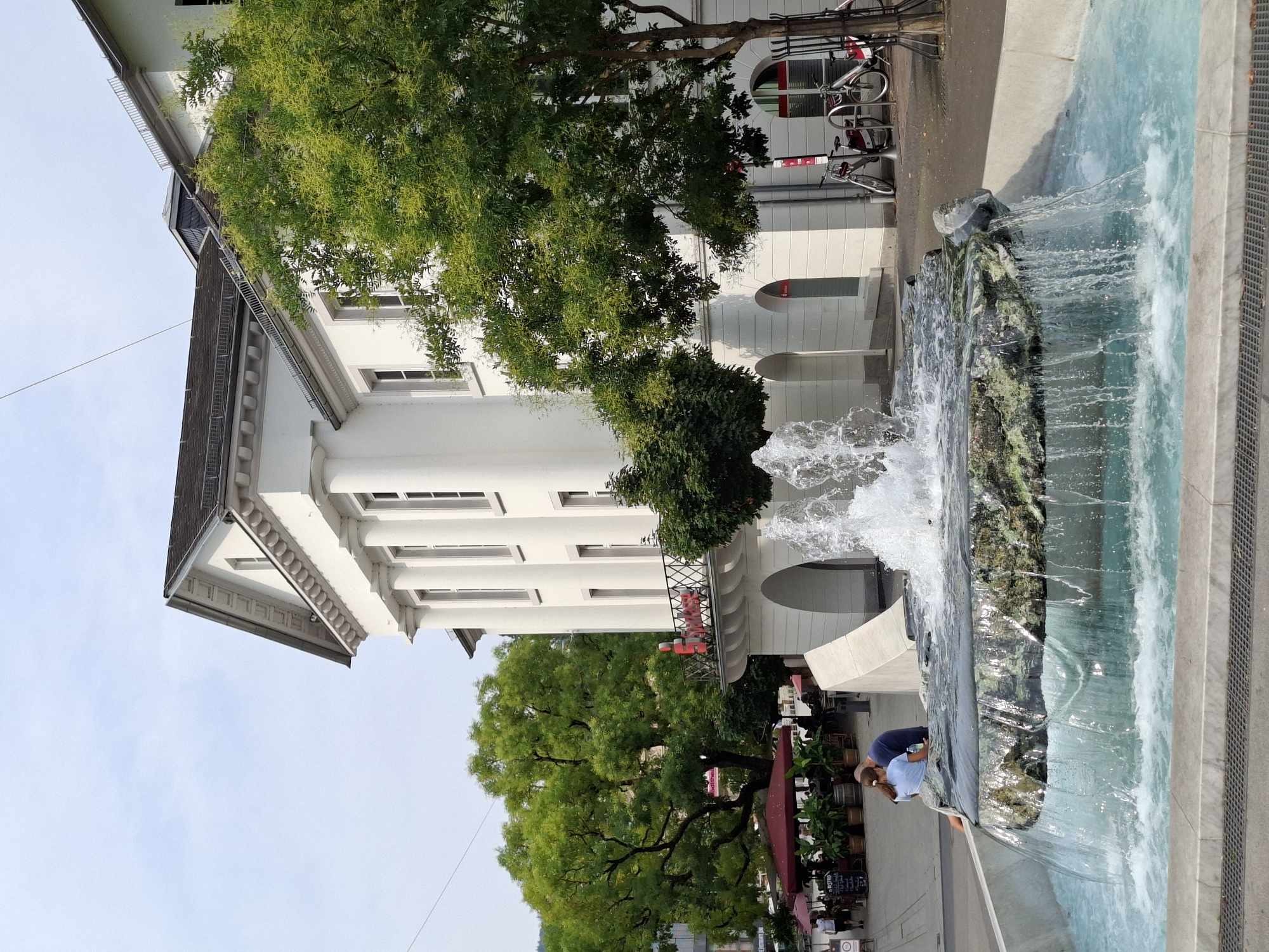An exterior, medium shot captures a public fountain in the foreground with a large white building and lush green trees in the background. The fountain features a rock formation with water cascading over its edges into a pool below. To the left of the fountain, a couple is seated, facing the water. The building behind the fountain is multi-story with a classical architectural style, including columns and arched doorways. Red umbrellas and outdoor seating are visible in the distance, suggesting a cafe or restaurant area. Several bicycles are parked near the building. The sky is light blue with some haze.