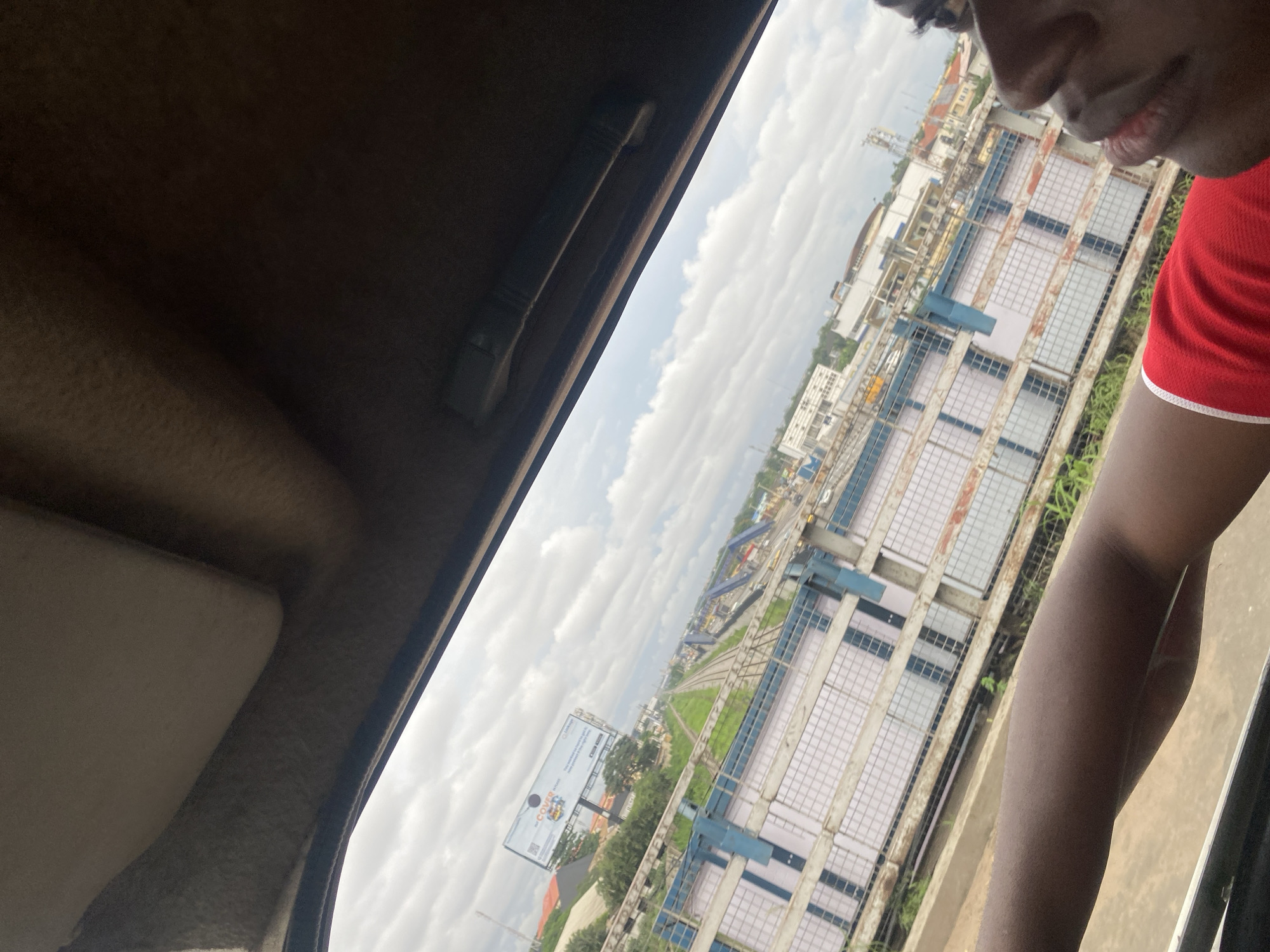 The image, taken from inside a vehicle looking out a window, captures a view of a cityscape, likely in Ikeja, Nigeria. The foreground is dominated by the arm of a person wearing a red shirt with white trim. The middle ground features a long, white and blue striped building with a metal railing in front of it. Beyond this building, a set of train tracks runs into the distance, flanked by greenery and more buildings. A large billboard displaying advertisements is visible in the upper left portion of the frame. The sky is partly cloudy. The scene suggests a movement-based perspective, possibly from a car or train traveling through an urban environment.