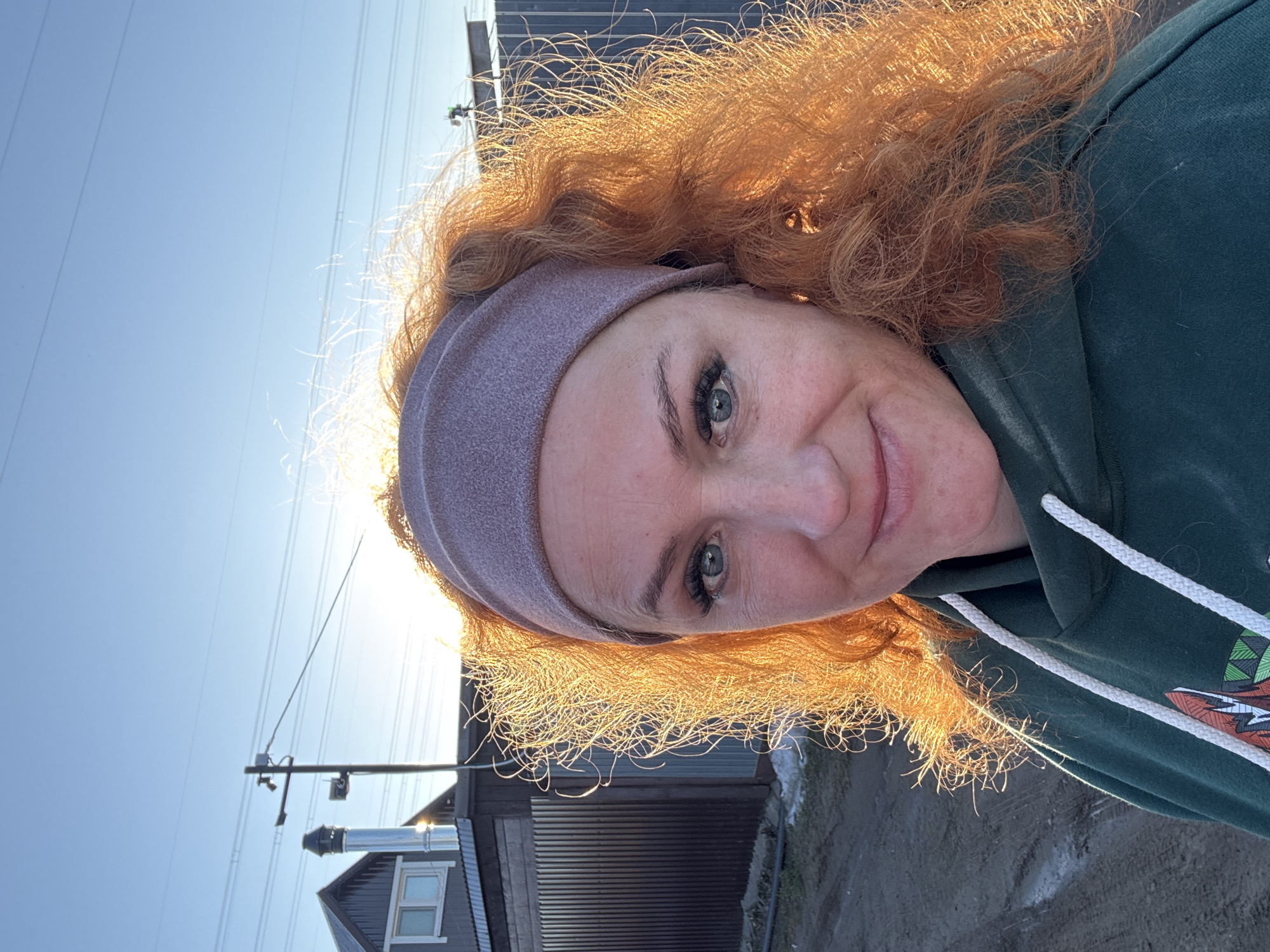 A woman with red curly hair, wearing a grey headband and a dark green hoodie, is taking a selfie outdoors. The background features a blue sky, a utility pole with wires, and a corrugated metal fence with a partial view of a building with a chimney. The scene appears to be located in a residential area. The location is determined to be Chaikovsky, Russia based on the presence of utility poles and the architecture of the buildings.