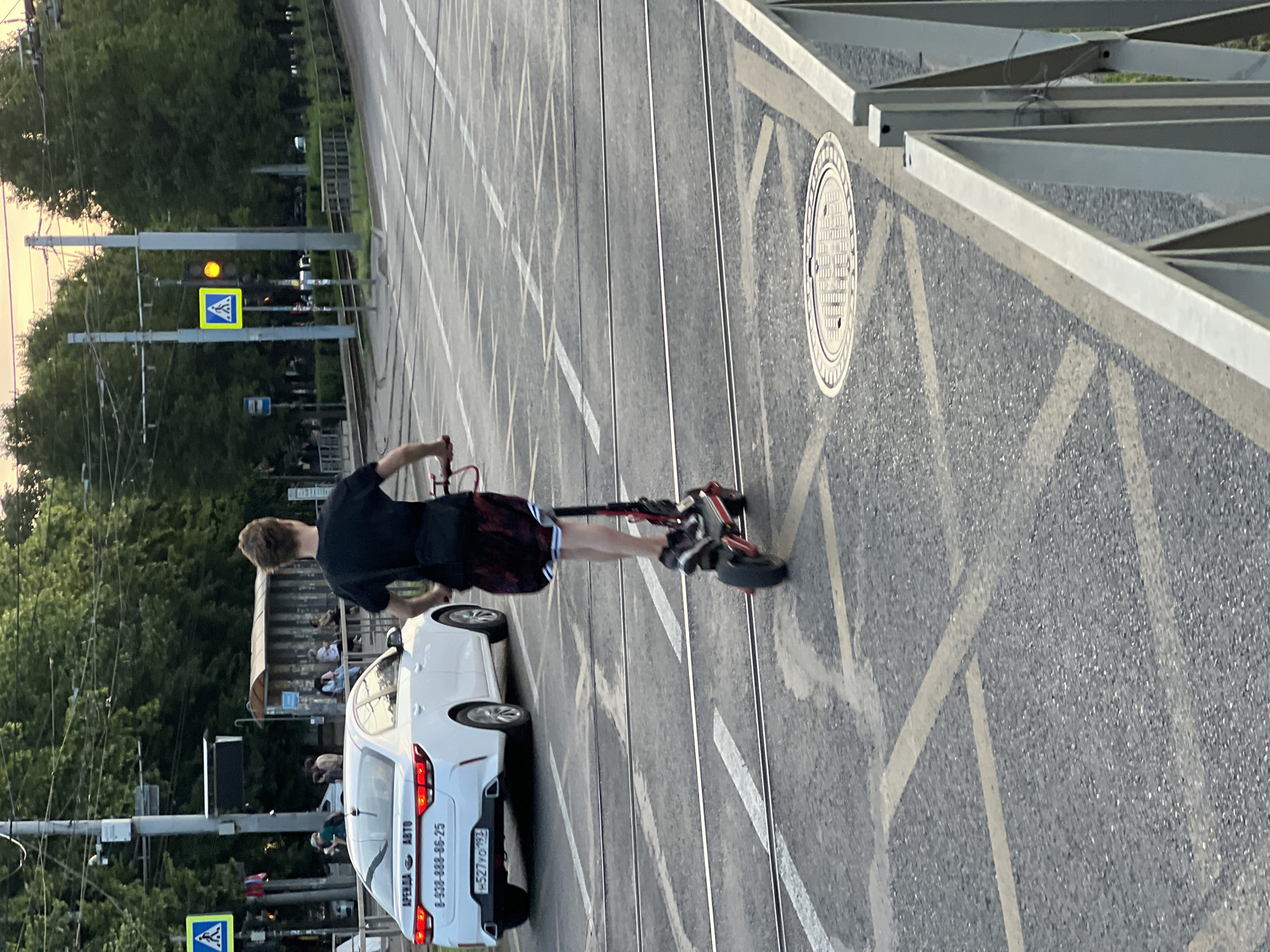 The image shows a young person riding an electric scooter on a city street in Krasnodar, Russia. The person is wearing a black t-shirt and patterned shorts. They are on a road with tram tracks. A white Kia car is parked on the left side of the road, with a license plate that reads "H527YO 193". There are pedestrian crossings marked on the road. In the background, there is a bus stop with people waiting. Trees line the street, and there are power lines overhead. The sky is a pale orange, suggesting it is either sunrise or sunset.