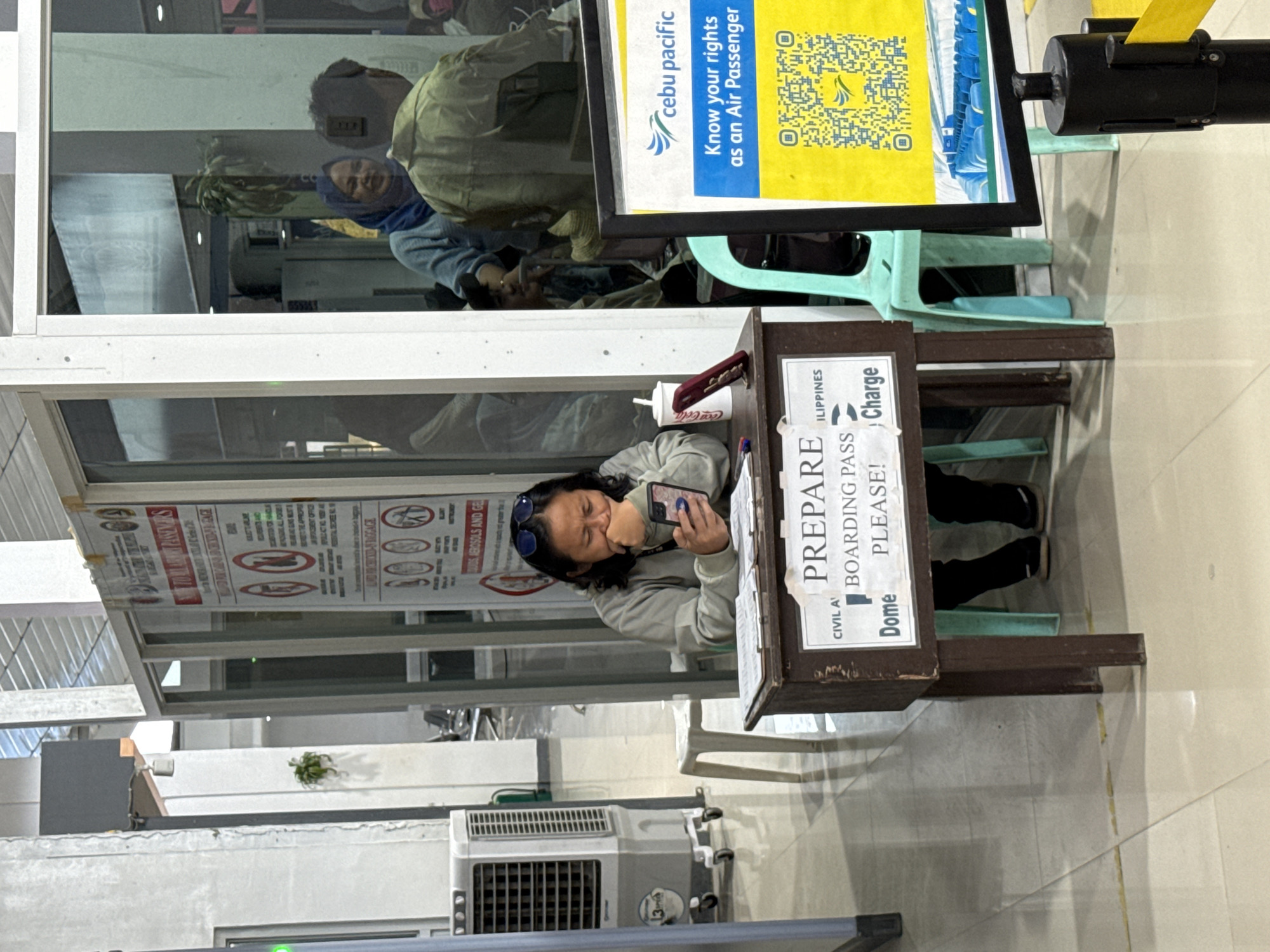 The image shows an indoor scene at an airport in the Philippines, likely in or near Del Carmen, based on the "ILIPPINES" text on the sign. A woman stands at a podium labeled "PREPARE BOARDING PASS PLEASE!" examining her phone. A cup of Coca-Cola sits on the podium near a pen. A sign advertising Cebu Pacific and air passenger rights is visible in the background. People are reflected in a window behind her. An air conditioning unit is located on the floor. Signs are posted with safety and prohibited items.