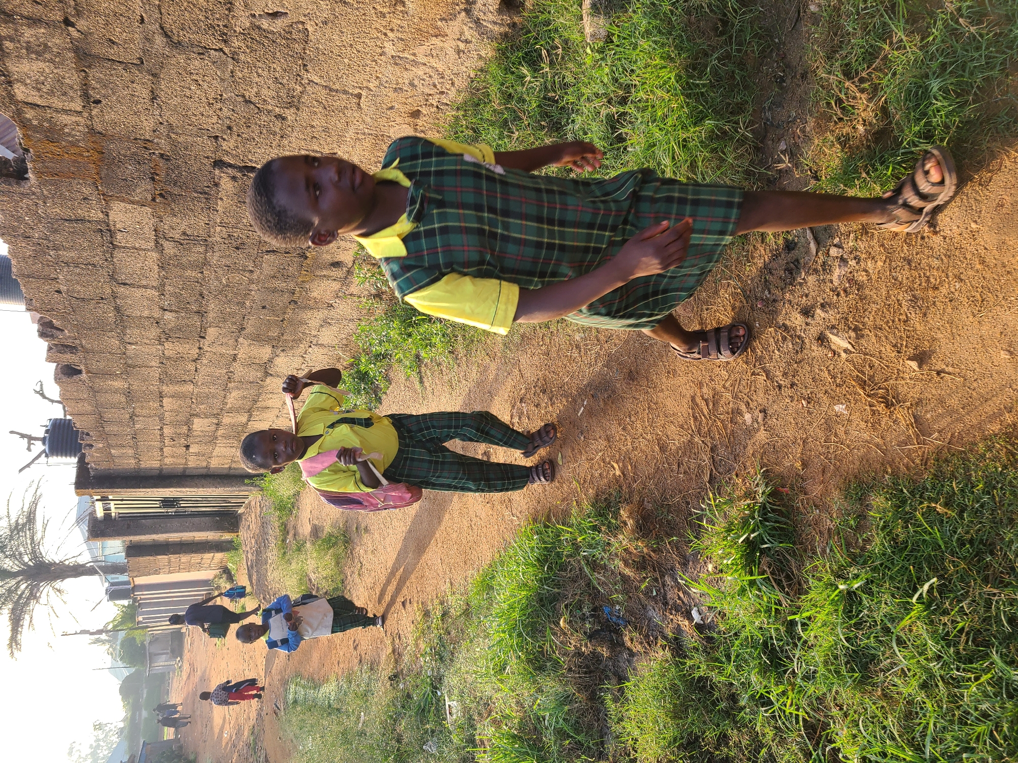 In Barakin Lerawi, Nigeria, a group of children walks along a dirt path beside a rough stone wall. The child in the foreground, on the right, wears a yellow shirt and a green and yellow plaid dress, with a pink backpack slung over their shoulders. They are wearing brown sandals and are walking towards the right of the frame. Behind them, another child in a similar uniform walks towards the left, also carrying a pink backpack. Further in the distance, more children are visible walking on the path, some with backpacks. The environment is rural, with sparse vegetation and a water tank visible on top of the wall in the background. The light suggests it is late afternoon.