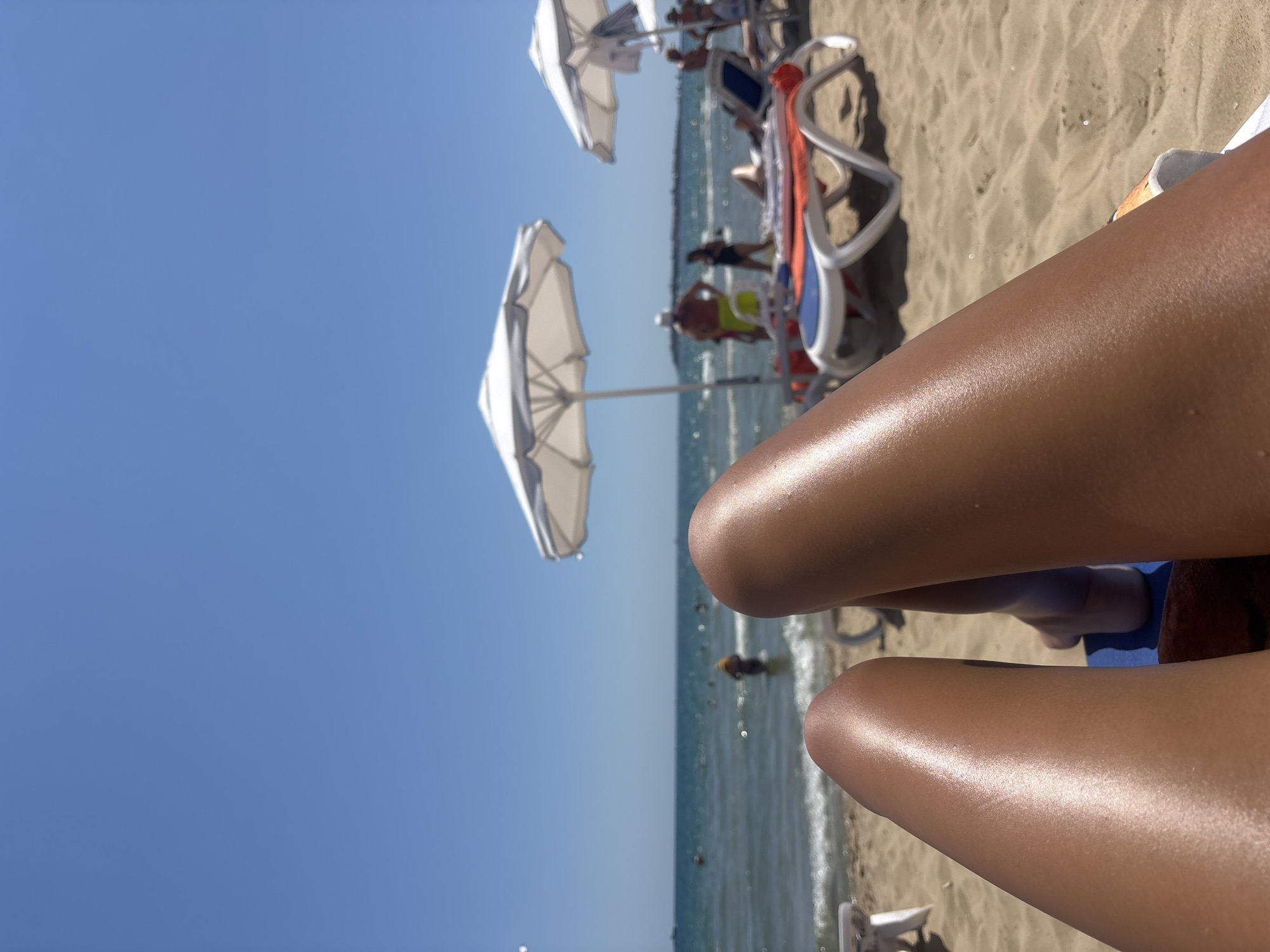 The image is a first-person perspective of someone relaxing on a beach in Nesebar, Bulgaria. In the foreground, the sun-tanned legs of the individual are visible, resting on a blue beach towel. The sand of the beach stretches out behind the legs. In the middle ground, several beach chairs and white umbrellas are set up. Some people are visible in the shallow water of the sea, while others are walking or sitting on the beach. The sea is calm and blue, and the sky is clear and bright. The overall impression is one of a sunny day at a popular tourist destination.