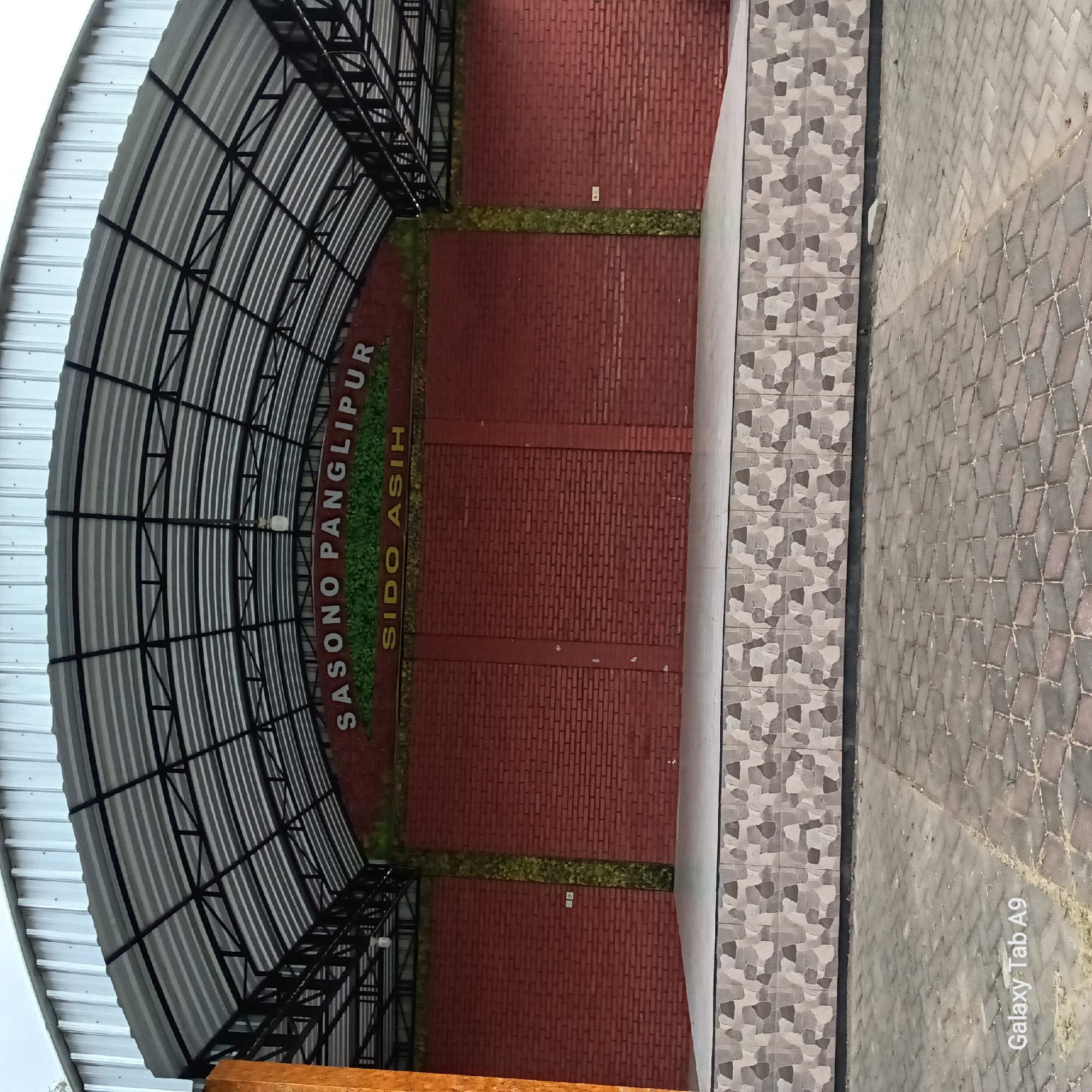 An outdoor stage area is depicted. The stage itself is elevated and has a tiled front with a gray, cream, and brown geometric pattern. The stage floor is a light gray. Behind the stage is a brick wall, painted red, with a vertical strip of green foliage on the left and right sides of the central section. Above the brick wall, large white lettering reads "SASONO PANGLIPUR" and below that, in yellow lettering, "SIDO ASIH". The stage is covered by a large, curved metal roof supported by a black metal frame. The roof is made of corrugated silver metal panels. The ground in front of the stage is paved with interlocking gray and pink rectangular bricks laid in a diamond pattern. The sky appears overcast. A white electrical outlet is visible on the red brick wall. The lower left corner of the image shows the text "Galaxy Tab A9".