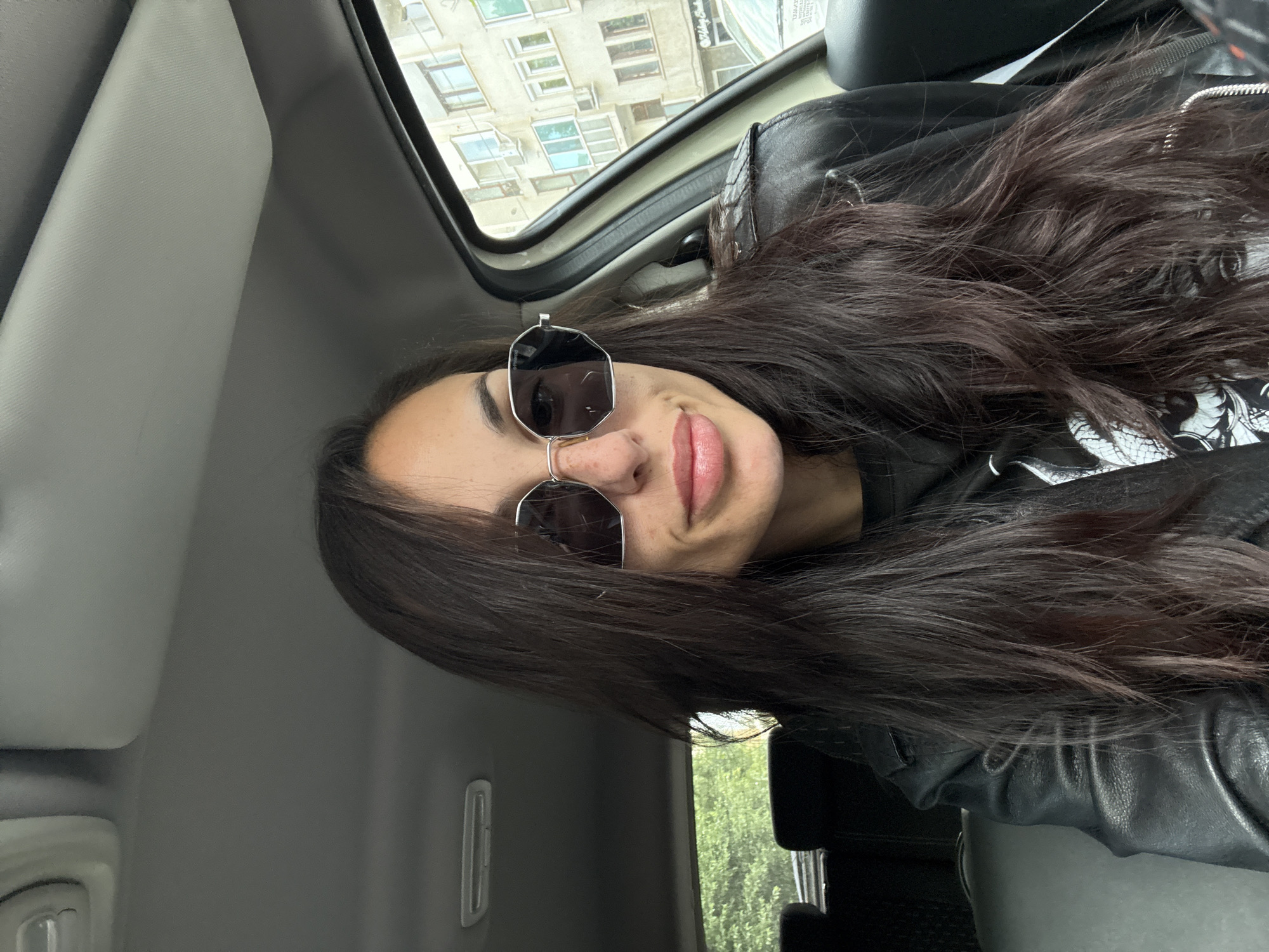 A woman with long, dark hair is wearing sunglasses while sitting in a car. The camera angle suggests the photo was taken from within the vehicle. Outside, a building with multiple windows is visible. The image does not contain information on the location in the city of Pleven, Bulgaria.