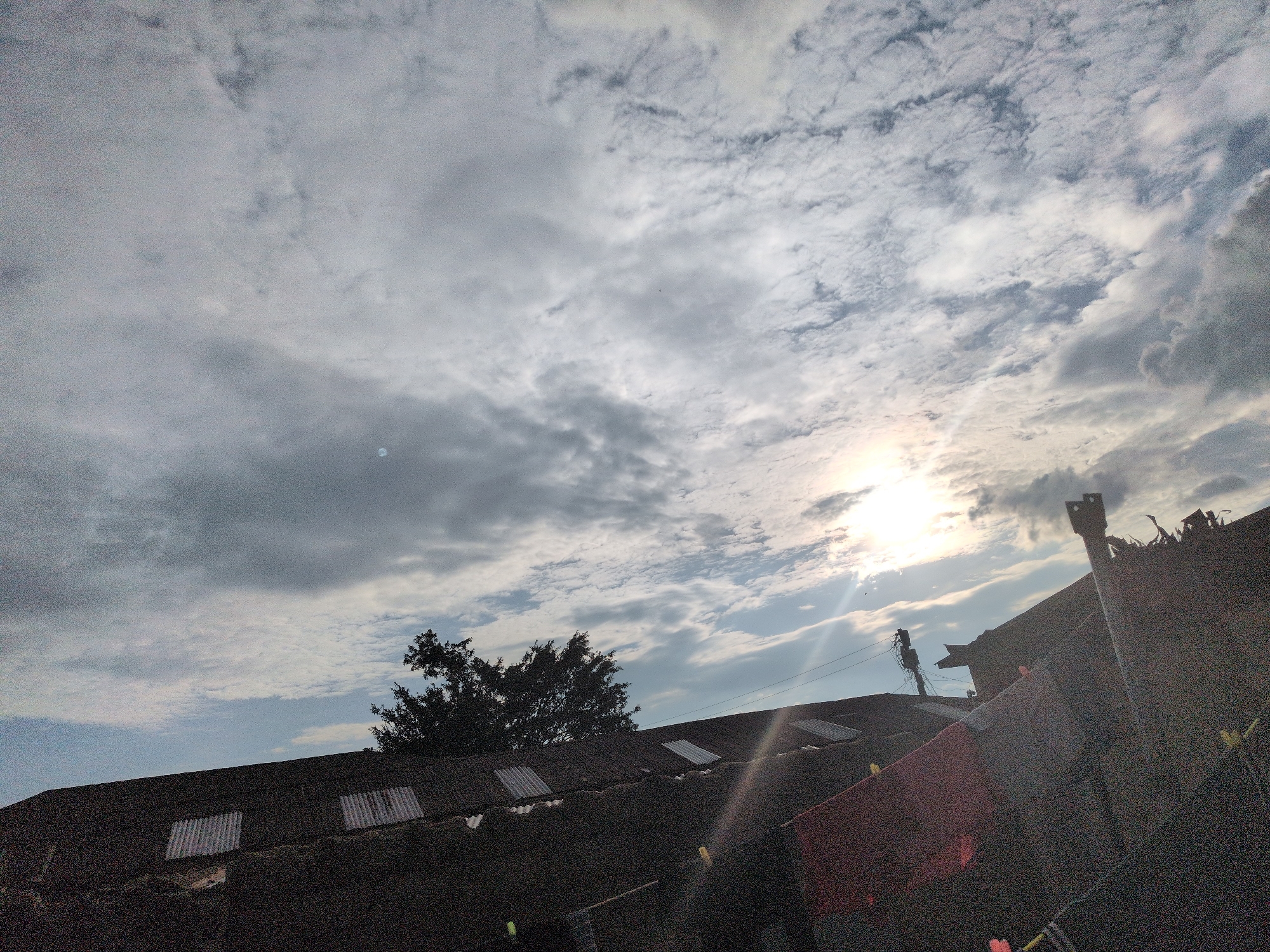 The image presents an outdoor scene captured in Jos, Nigeria, characterized by a bright, partly cloudy sky above silhouetted rooftops and vegetation. The upper two-thirds of the frame are dominated by a sky featuring a mixture of scattered white and grey cumulus and stratocumulus clouds, with occasional patches of clear, light blue sky visible. A bright, low-angle sun is prominently positioned in the upper right quadrant, partially obscured by clouds, generating a distinct lens flare that radiates downwards and leftwards across the sky. Two small, circular, bright white spots are discernible in the sky; one is in the upper left, and another is to the lower right of the main sun flare, closer to the horizon.
Below the sky, the lower third of the image is occupied by several dark, sloping corrugated iron rooftops, heavily shadowed and appearing almost as silhouettes due to the backlighting from the sun. These roofs exhibit lighter, possibly translucent or reflective, rectangular sections. A dense mass of dark green foliage, indicative of trees, rises behind the rooftops on the left side, appearing as a strong silhouette against the brighter sky. On the right side, extending above the rooftops, are dark, angular structures, including what appears to be a chimney or vent, and another structure resembling a utility pole with visible crossarms and wires. On one of the foreground rooftops, laundry hangs on a clothesline, secured by bright yellow pegs to dark-colored fabric. No people are visibly present.