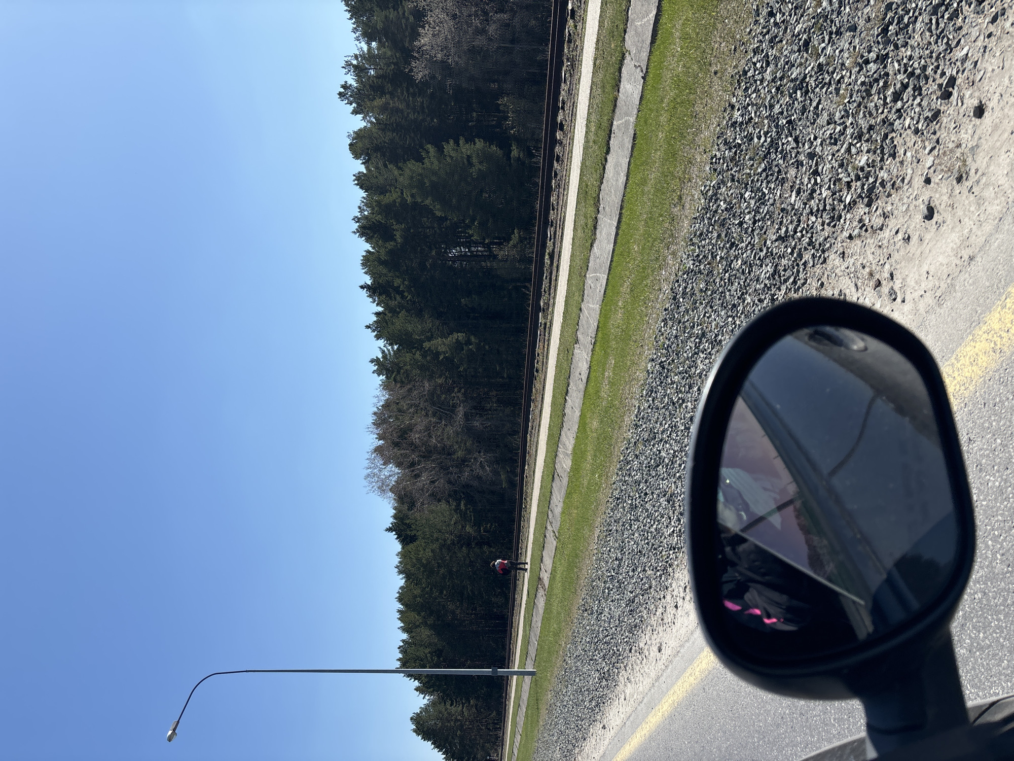 The image shows a view from a vehicle's side mirror. The location is in Chaikovsky, Russia, evidenced by the presence of a Russian railroad. The environment features a clear blue sky, a dense coniferous forest, and a train track. A person wearing a red jacket is standing near the tracks. The immediate foreground consists of a paved road with a yellow line and gravel on the side. The side mirror reflects the interior of the vehicle.