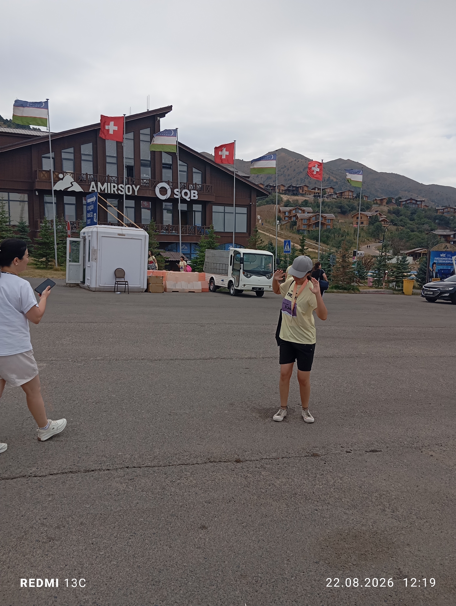 In the city of Amirsay, Uzbekistan, a ski resort is depicted under a cloudy sky. The main building of the resort features a wooden facade with large windows and the prominent sign "AMIRSOY SQB." Flags of Uzbekistan and Switzerland are displayed in front of the building.
In the foreground, a woman in a white t-shirt and beige shorts holds a smartphone and walks away from the camera, while another woman, wearing a yellow t-shirt, black shorts, and a grey cap, stands facing the camera with her hands raised. She is wearing a race bib with the number 536.
Several vehicles are present in the scene, including a white shuttle bus and a black car parked on the right. A white security booth with a chair inside is located near the entrance of the resort. Orange traffic barriers are also visible. The background shows a hillside dotted with numerous houses and trees, typical of a mountain resort. The overall environment suggests a recreational or sporting event is taking place.