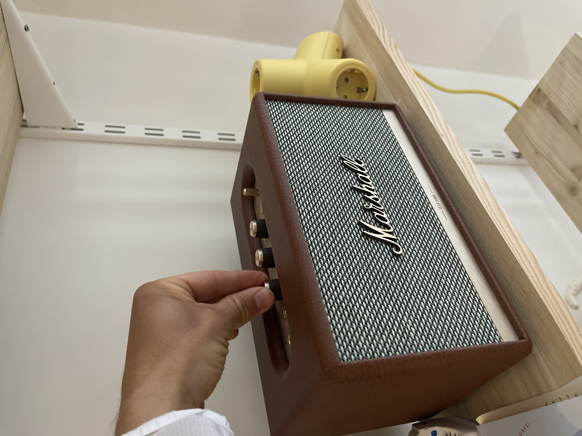 A person's hand is manipulating a knob on a Marshall Acton II Bluetooth speaker, which sits on a shelf. A yellow power adapter is plugged into a socket above the speaker. The shelf is white with metal tracks and wooden supports. The scene is lit by natural light. There is no indication of the location being Izhevsk, Russia.