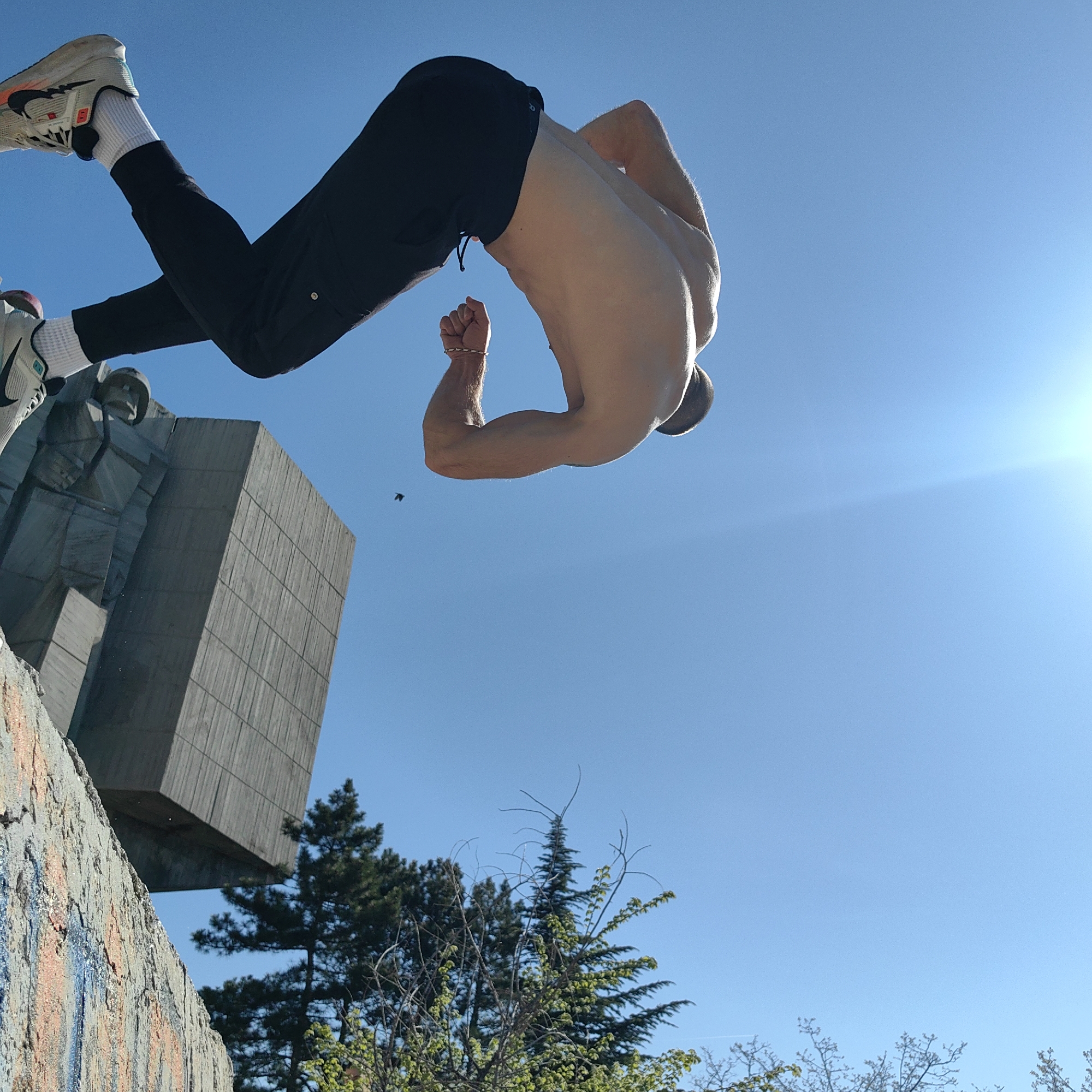 This dynamic outdoor image captures a shirtless male athlete in mid-air, performing an acrobatic flip or stunt against a brilliant, clear blue sky. He is inverted, with his body bent, wearing black athletic trousers and white Nike sneakers with prominent orange and black details. One of his hands is clenched into a fist, showcasing his muscular physique.
The setting features a large, brutalist-style concrete monument or building, composed of stacked geometric blocks, visible to the left. This architectural style, combined with the Varna, Bulgaria location hint, strongly suggests the Monument to the Bulgarian-Soviet Friendship, also known as the Pantheon. Below and around this structure, green trees and foliage are visible. The bottom-left corner shows a textured, possibly graffitied, wall or ledge, from which the athlete likely initiated his jump.
The time of day is bright and sunny, indicated by the vibrant blue sky and a distinct sun flare in the upper right quadrant. A tiny, dark object, possibly a bird or insect, can be seen flying in the sky near the concrete edifice. The image conveys a sense of athleticism, agility, and the freedom of movement often associated with parkour or gymnastics.