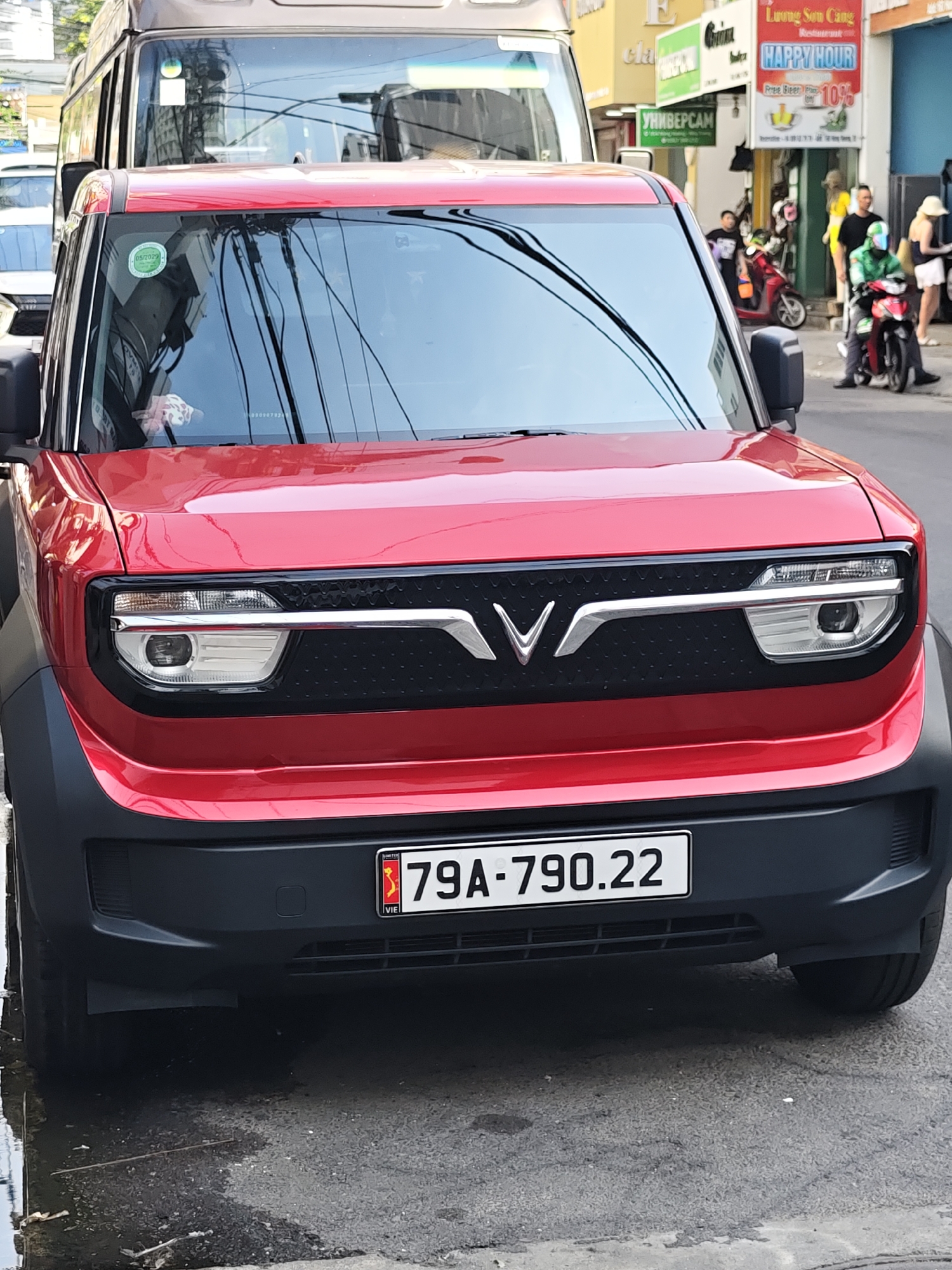 A red VinFast VF e34 electric car is parked on a street in Nha Trang Ward, Vietnam. The car has a black grille with a silver V-shaped logo, and its headlights are visible. A license plate reading "79A-790.22" is affixed to the front of the car. The car is positioned in front of a brown van. In the background, several people and motorbikes are visible. A "HAPPY HOUR" sign with a 10% discount is displayed on a building. The street is paved and appears wet in some areas, suggesting recent rain. Overhead wires are strung across the scene.