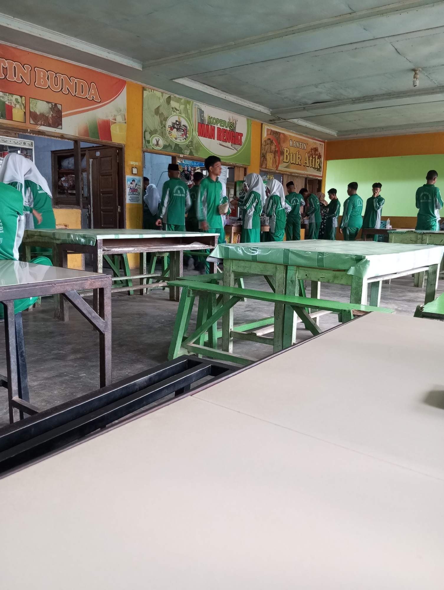 The image depicts an indoor scene, likely a school cafeteria or common area, in Pematang Reba, Indonesia. The space is furnished with several long tables, some covered with green plastic sheeting, and benches. In the background, murals and signage are visible on the walls, including one that reads "TIN BUNDA" and another with "KANTIN Buk Atik."
A group of young people, predominantly wearing green uniforms with white accents and headscarves for some, are gathered. Some are standing and interacting, while others appear to be in transit. The overall atmosphere suggests a casual, possibly break-time, setting. The flooring is concrete, and the ceiling appears to be made of wood panels with exposed light fixtures. Natural light enters from the left, and there is also artificial lighting.